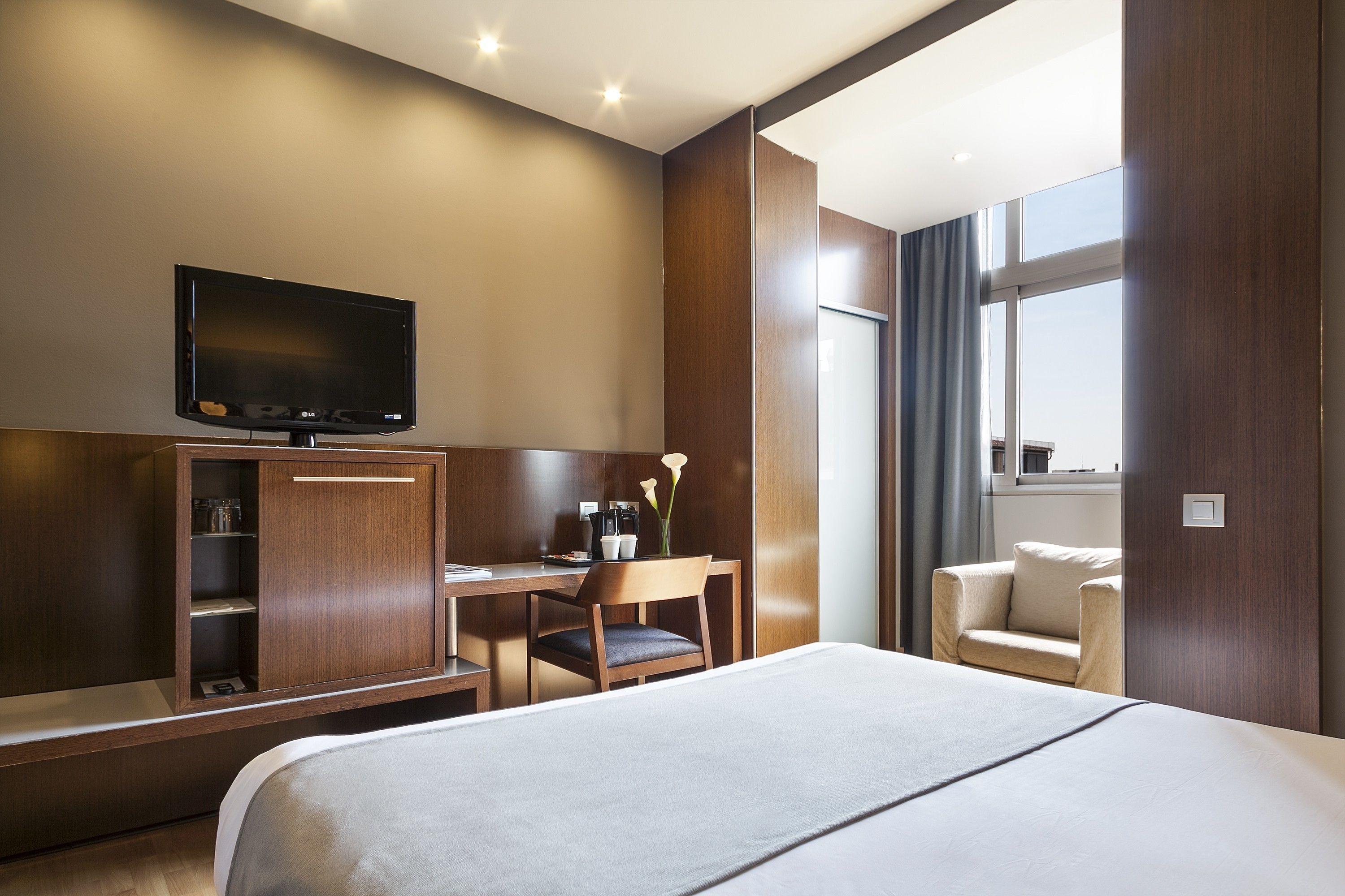 Habitación de hotel con cama, escritorio, silla, televisor y ventana con cortinas.