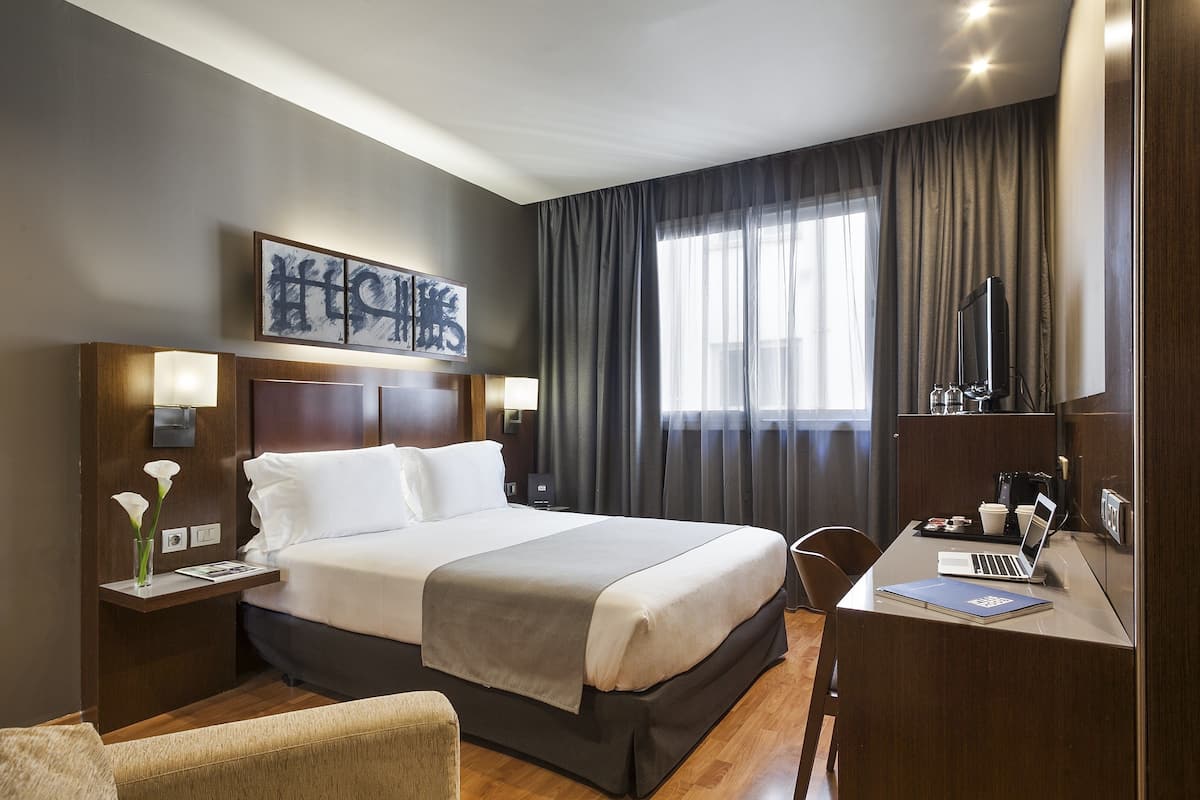 Habitación de hotel con una cama grande, un escritorio con una computadora portátil, una silla, una mesita de noche con una lámpara y una ventana con cortinas.
