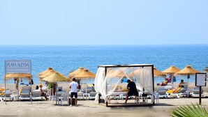 A beach with thatched umbrellas, lounge chairs, and a sign reading "PLAYA ALEYSA".