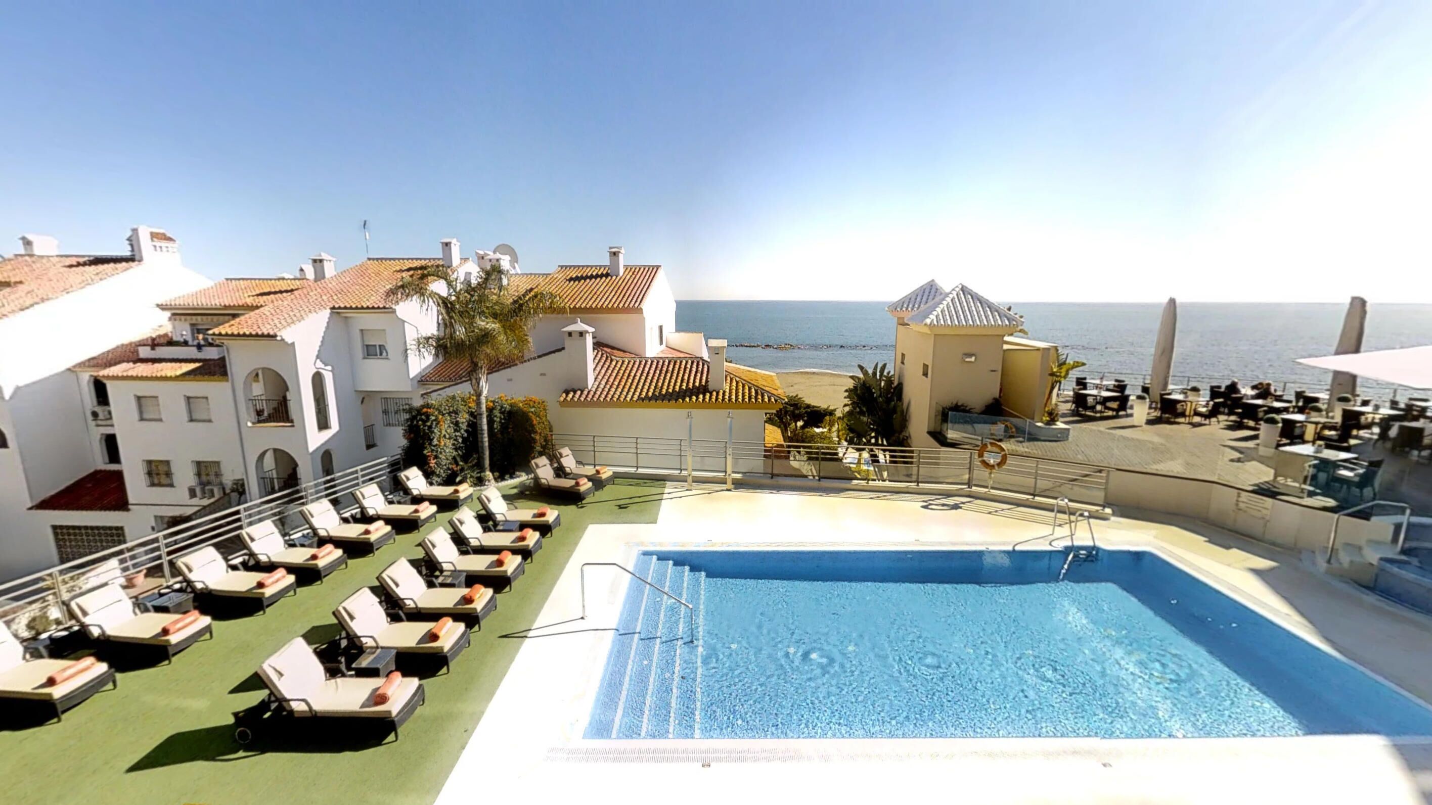 A rooftop pool with lounge chairs, a clear view of the sea, and a building with a tiled roof.