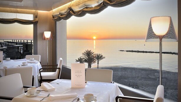 A dining area with tables set for breakfast, a menu stand, and a view of the sunset over the water.
