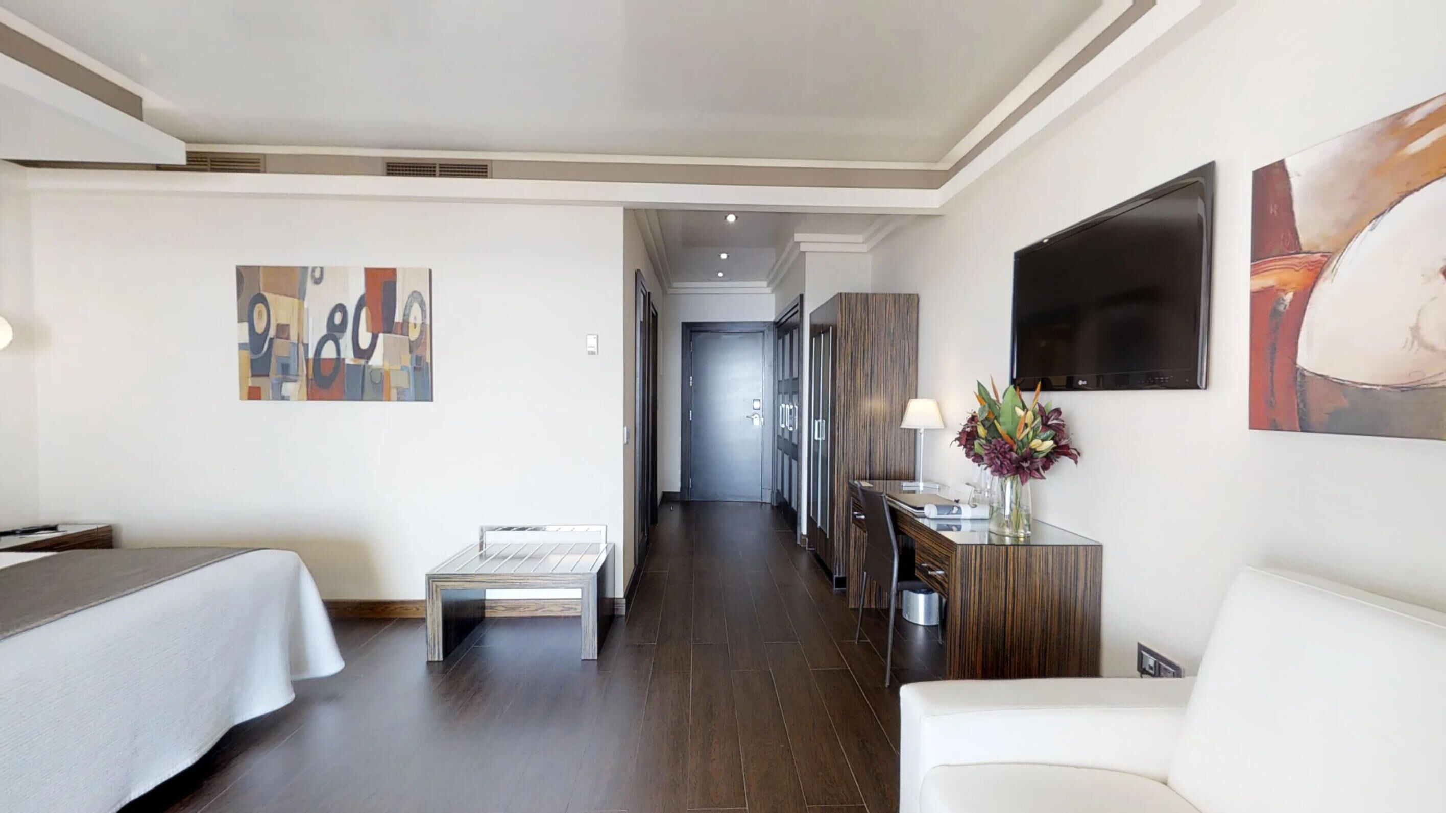 A hotel room with a bed, a desk with a lamp, a chair, and artwork on the walls.