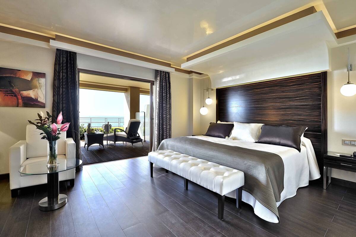 Ein modernes Hotelzimmer mit einem großen Bett, einem Glastisch mit Blumen, einer Bank, Blick ins Freie und einem dunklen Holz-Kopfteil.