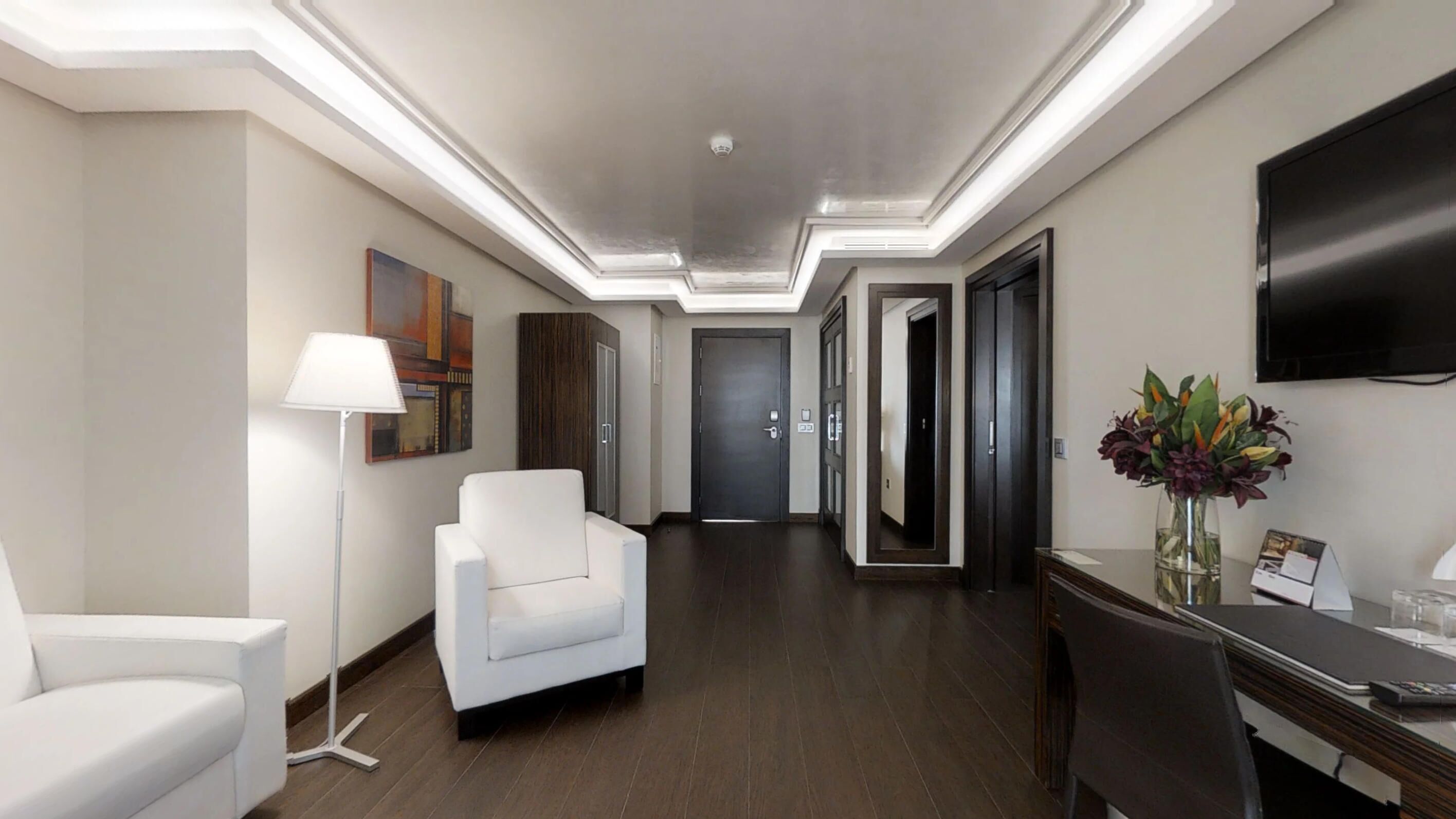 A hotel hallway with a white armchair, a desk with a vase of flowers, and a flat-screen TV on the wall.