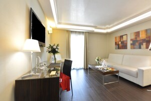 Ein modernes Hotelzimmer mit flachbildfernseher an der Wand, einem Essbereich mit Tisch und Stühlen, einem weißen Sofa und einem Couchtisch mit Blumen.