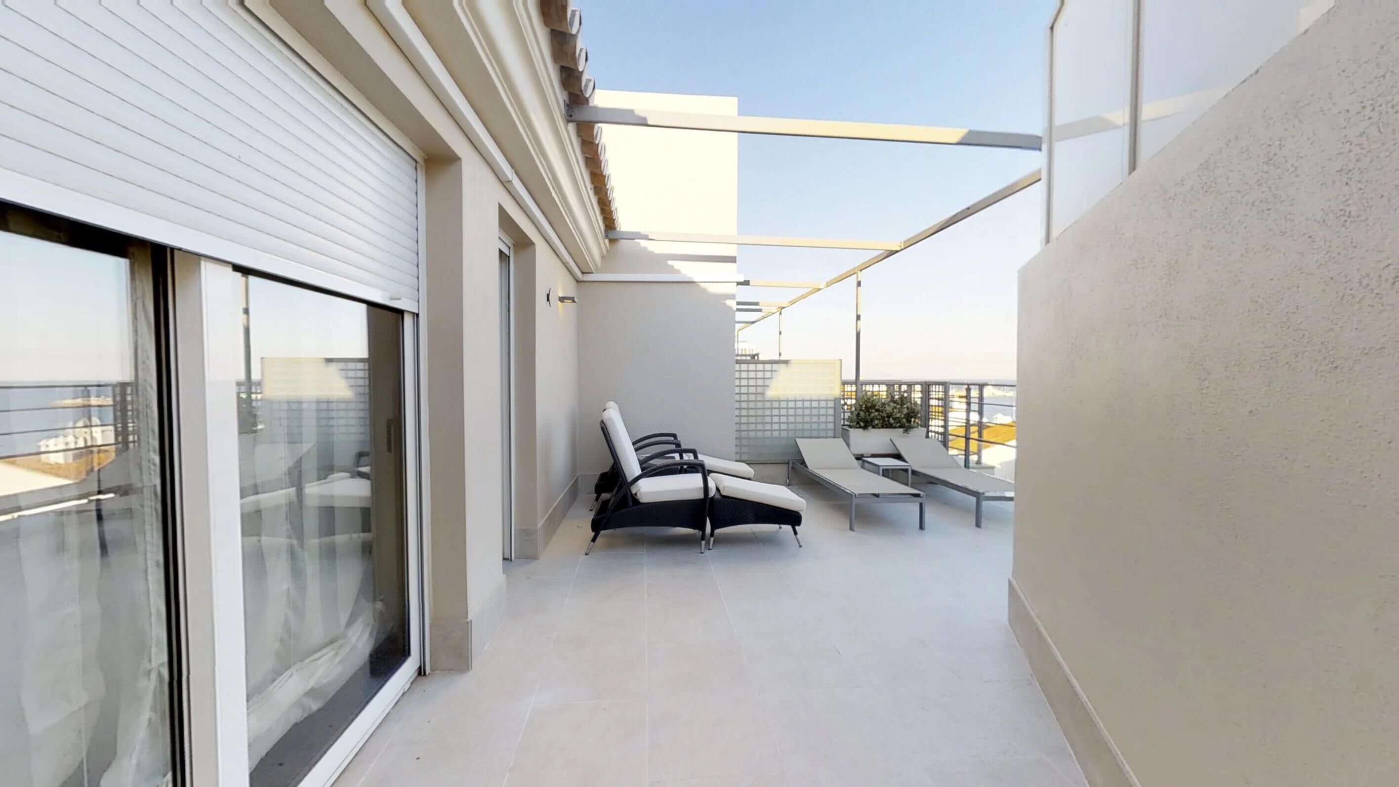 A modern balcony with a glass door, two lounge chairs, a small table, and a potted plant.