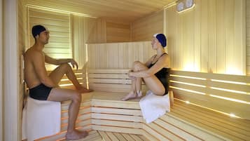 A sauna with wooden benches and a sloped floor, equipped with wall-mounted clocks.