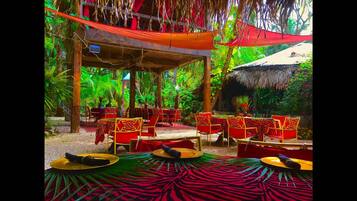 An outdoor seating area with red and gold chairs and tables, surrounded by lush greenery and a thatched-roof structure.