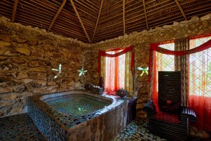 Una habitación con paredes de piedra, una bañera grande de azulejos y una silla de madera.