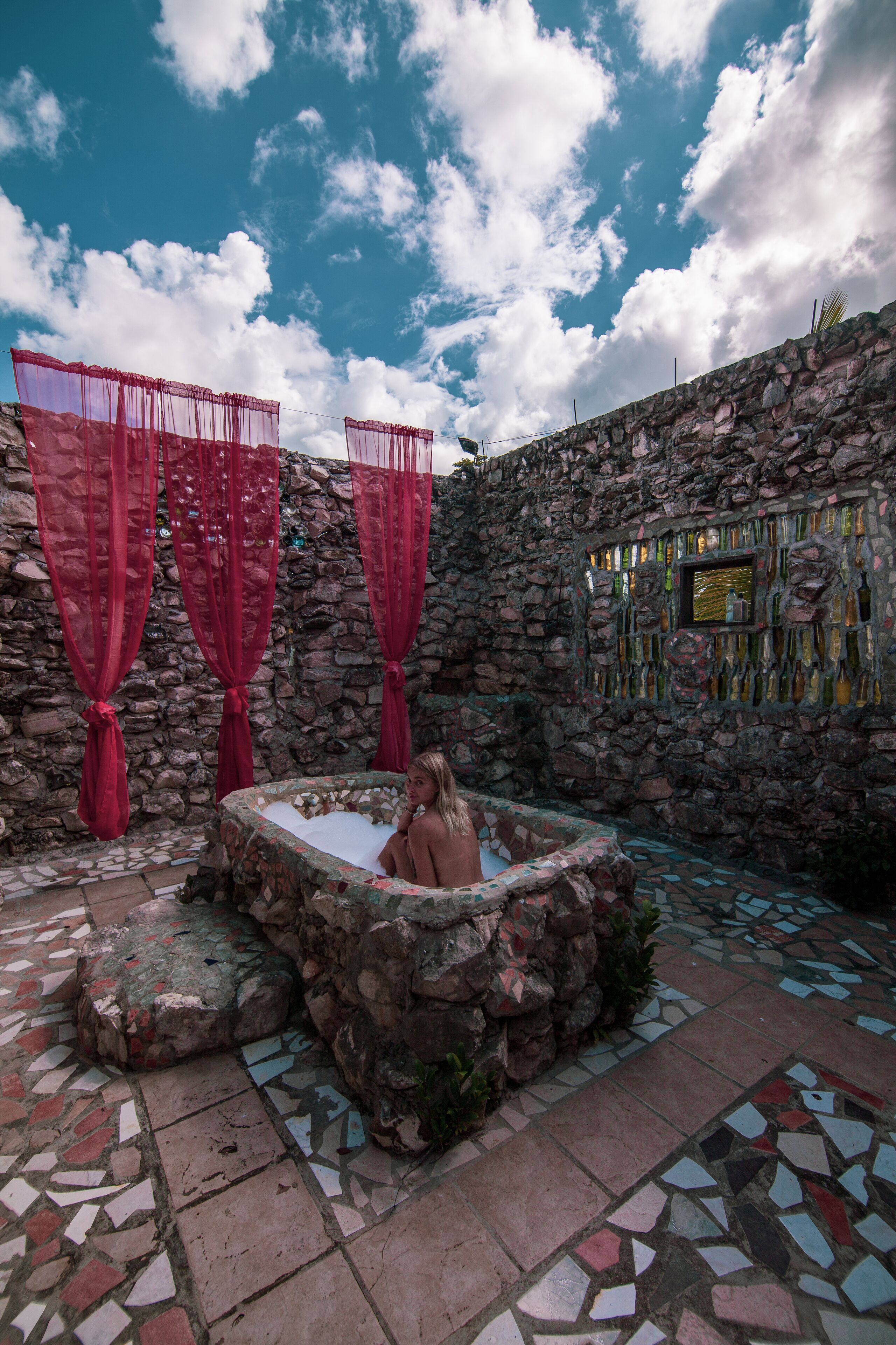 A stone-walled outdoor area with a mosaic-tiled floor, a hot tub, and red curtains.