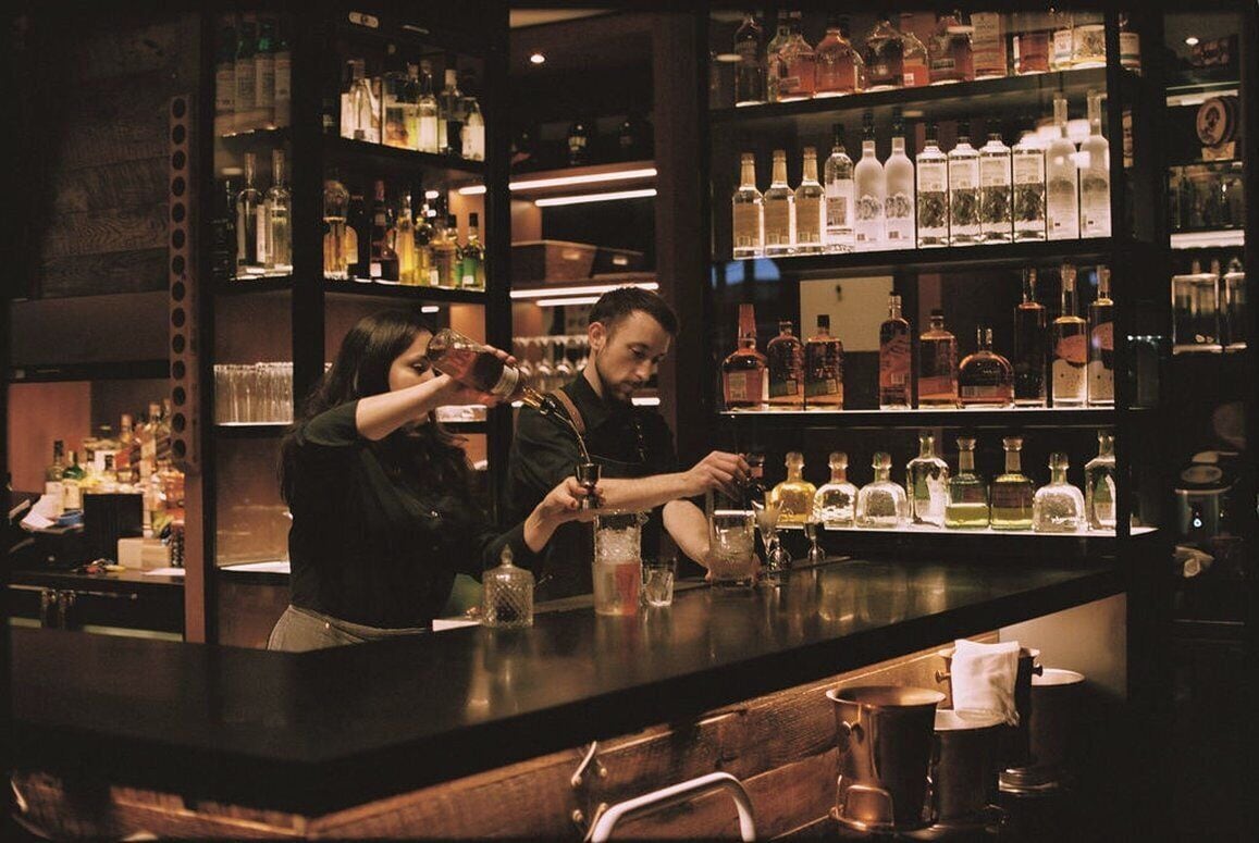 Un espace bar avec des étagères remplies de bouteilles d’alcool et un barman qui prépare des boissons.