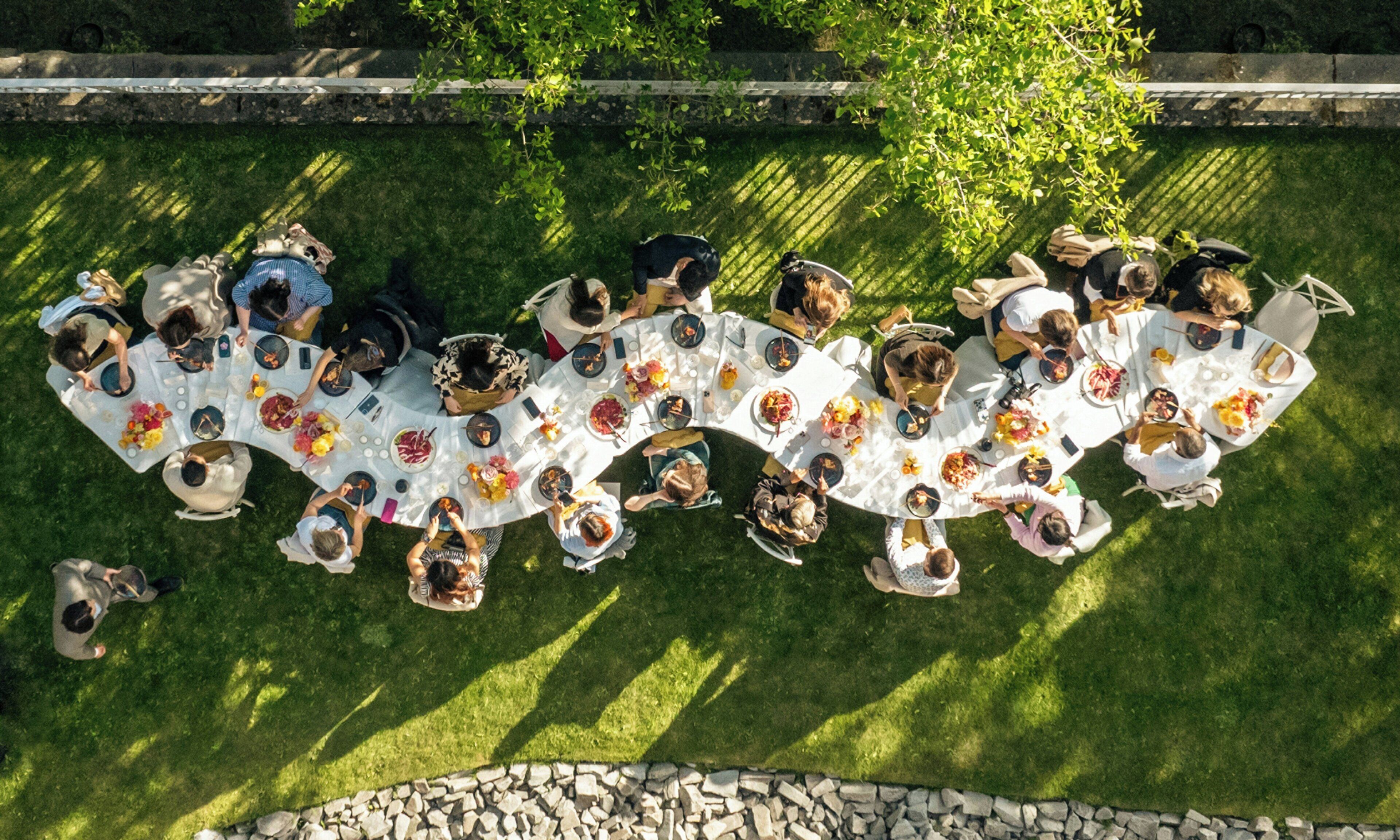 An aerial view of a group of people seated around a large, round table outdoors.