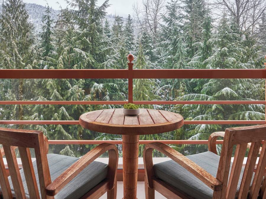 A wooden balcony with a round table and two chairs, overlooking a forested area.