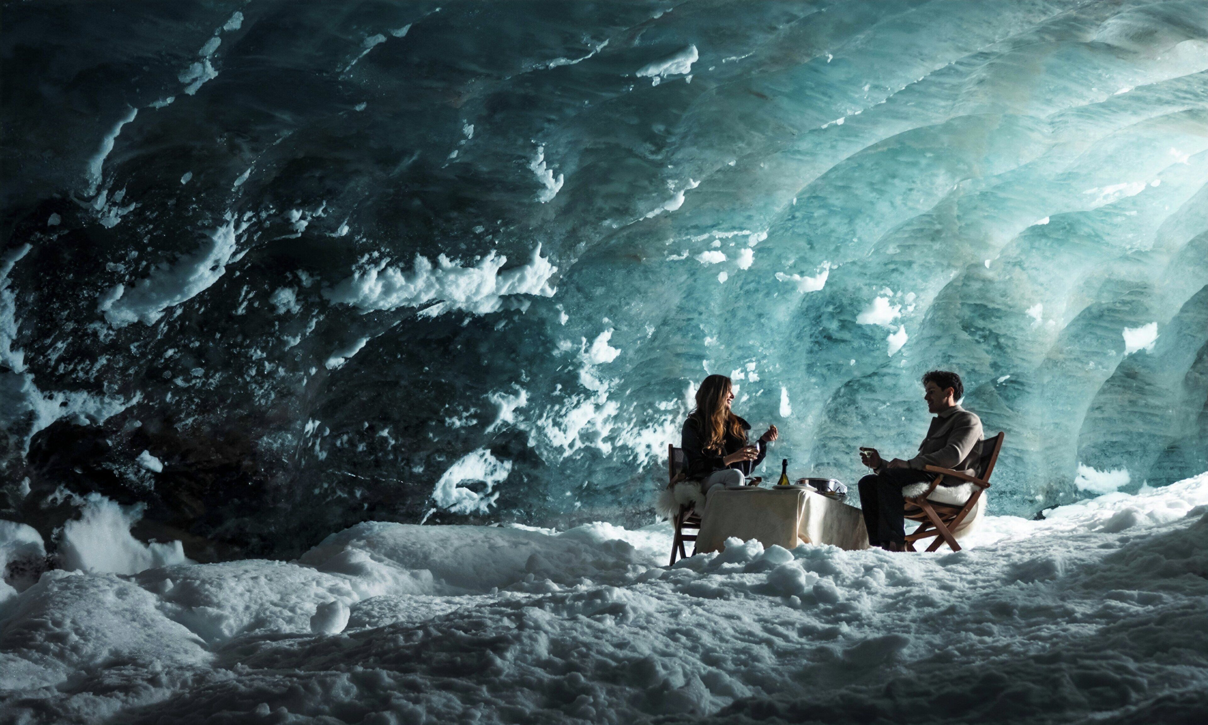 An ice cave interior with two people seated at a table, surrounded by snow.