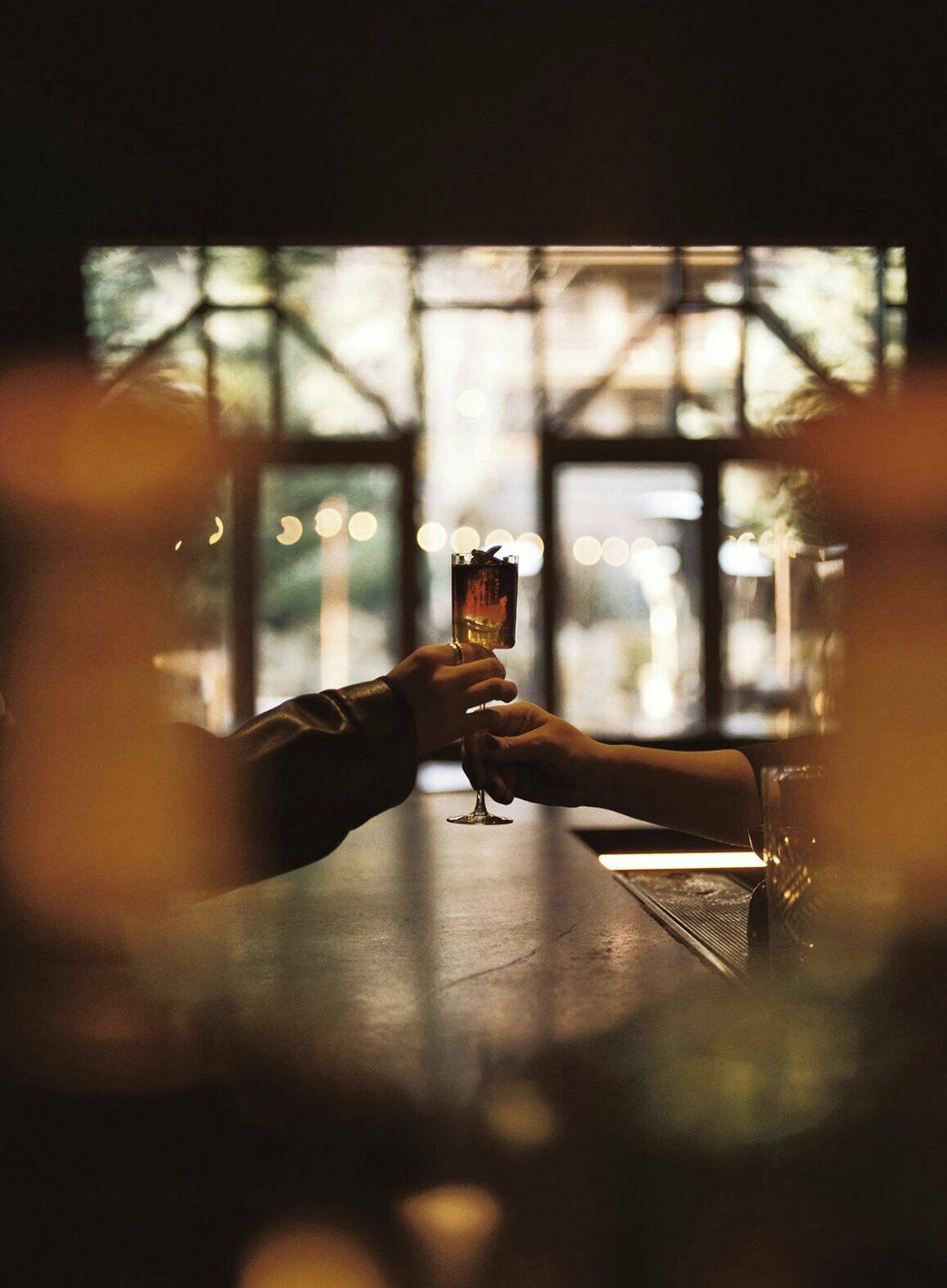 Two hands holding a glass of drink at a bar with a view through large windows.