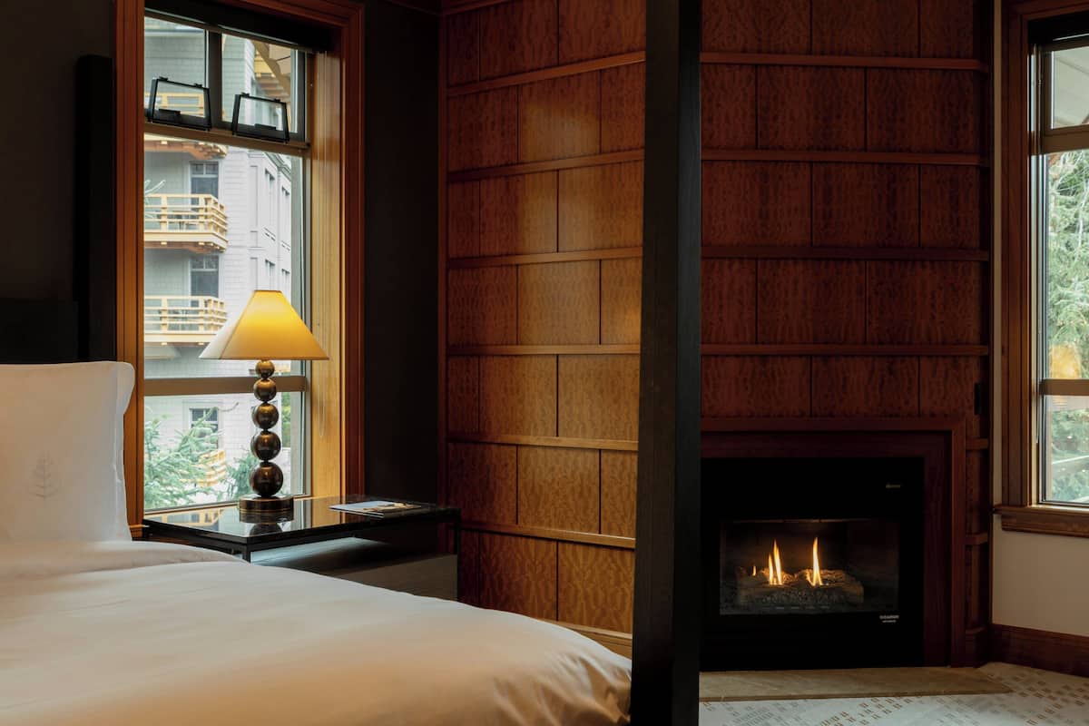 Habitación de hotel con cama, mesita de noche con lámpara, chimenea y ventana con vista a edificios.