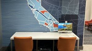 A hotel room with a large wall map, a desk with two chairs, and a computer monitor displaying a scenic landscape.