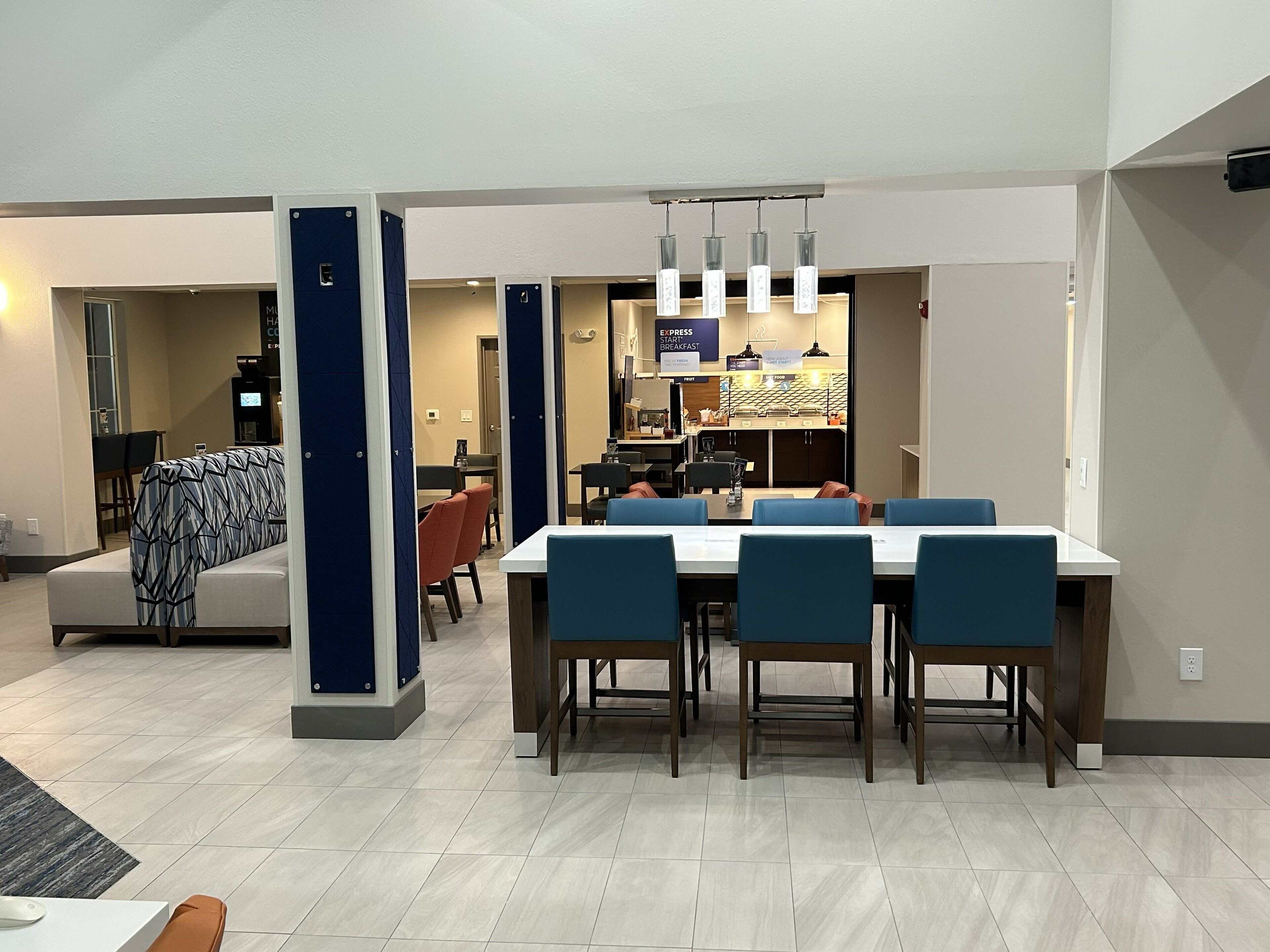 A modern dining area with a long table and blue chairs, a kitchen in the background, and a sofa with a patterned cover.