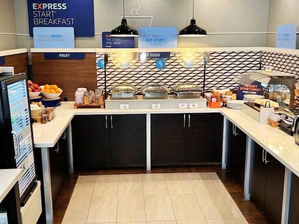 A breakfast buffet area with labeled food stations, a fruit section, and a sign asking about the start of the day.
