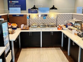 Zona de desayuno tipo buffet con estaciones de comida señalizadas, una sección de frutas y un cartel preguntando por el horario de inicio.