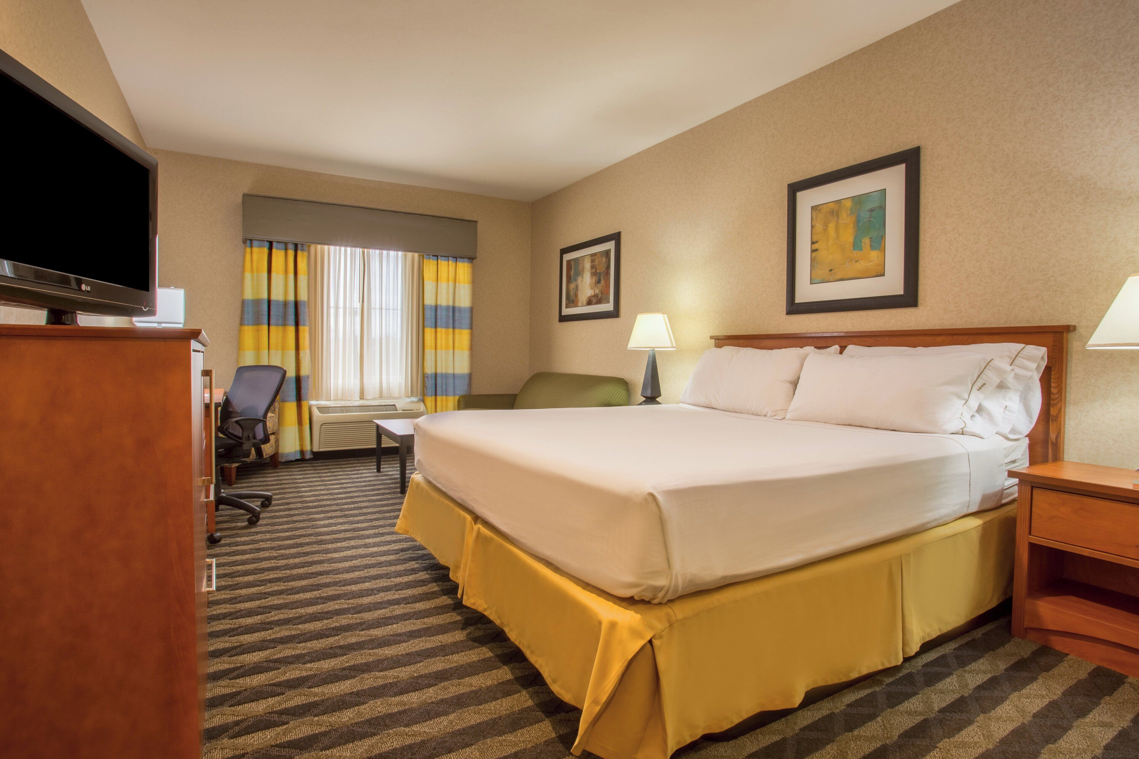 Quarto de hotel com cama, escrivaninha, cadeira, televisão e janela com cortinas.