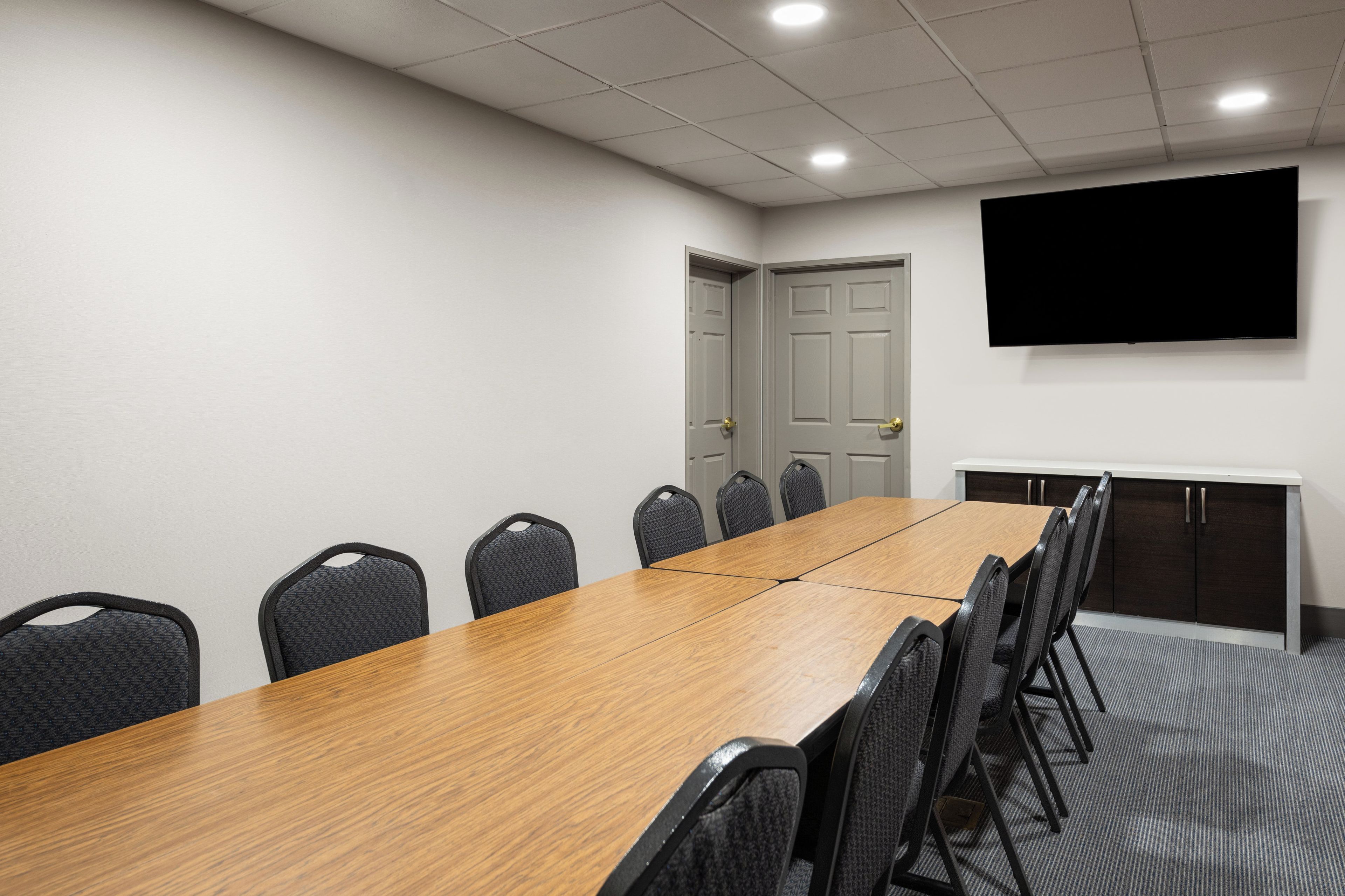 A conference room with a long wooden table, six chairs, a flat-screen TV on the wall, and a door.