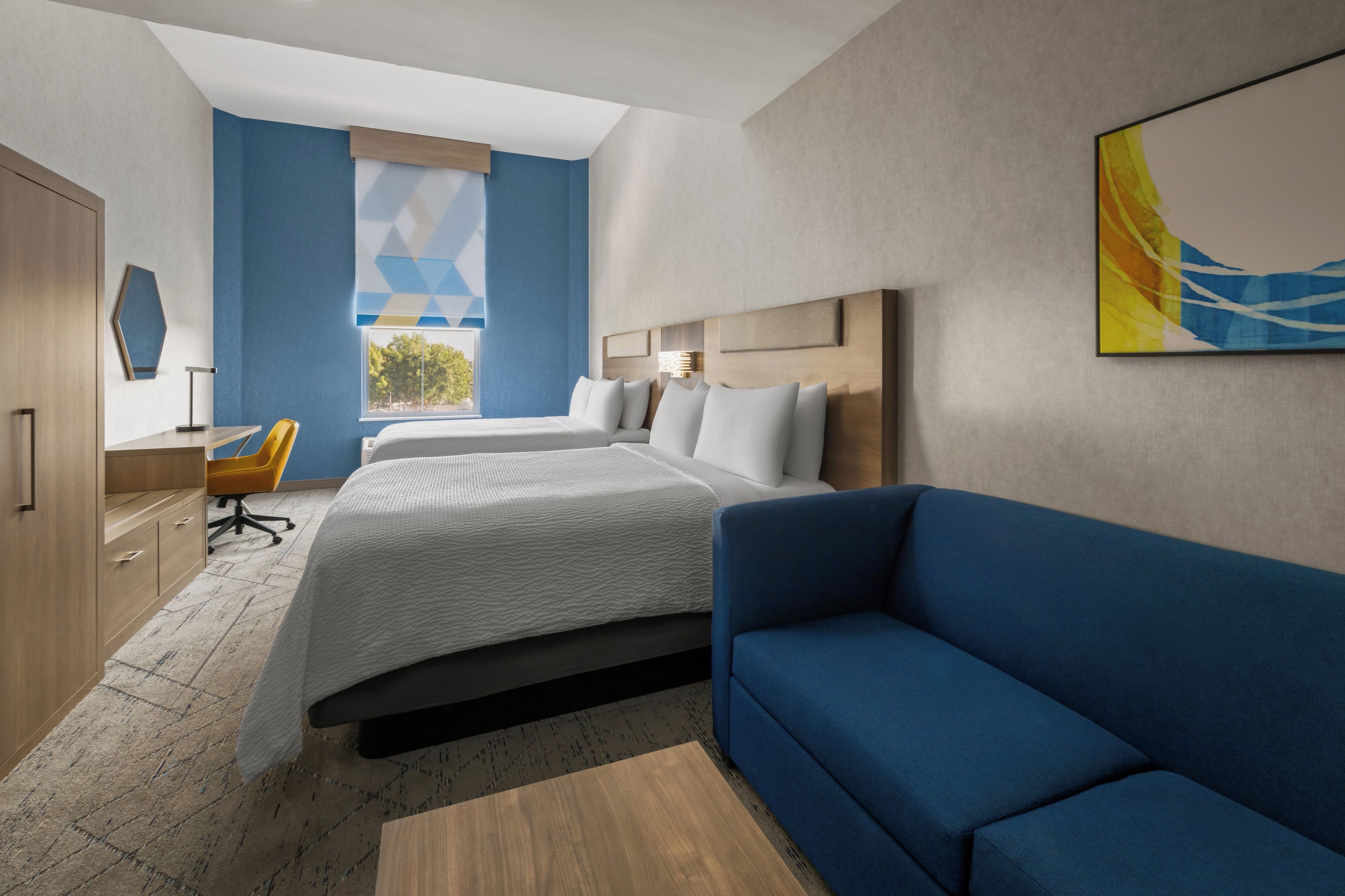 A modern hotel room with a large bed, a blue sofa, a desk with a chair, and a window with a view of trees.