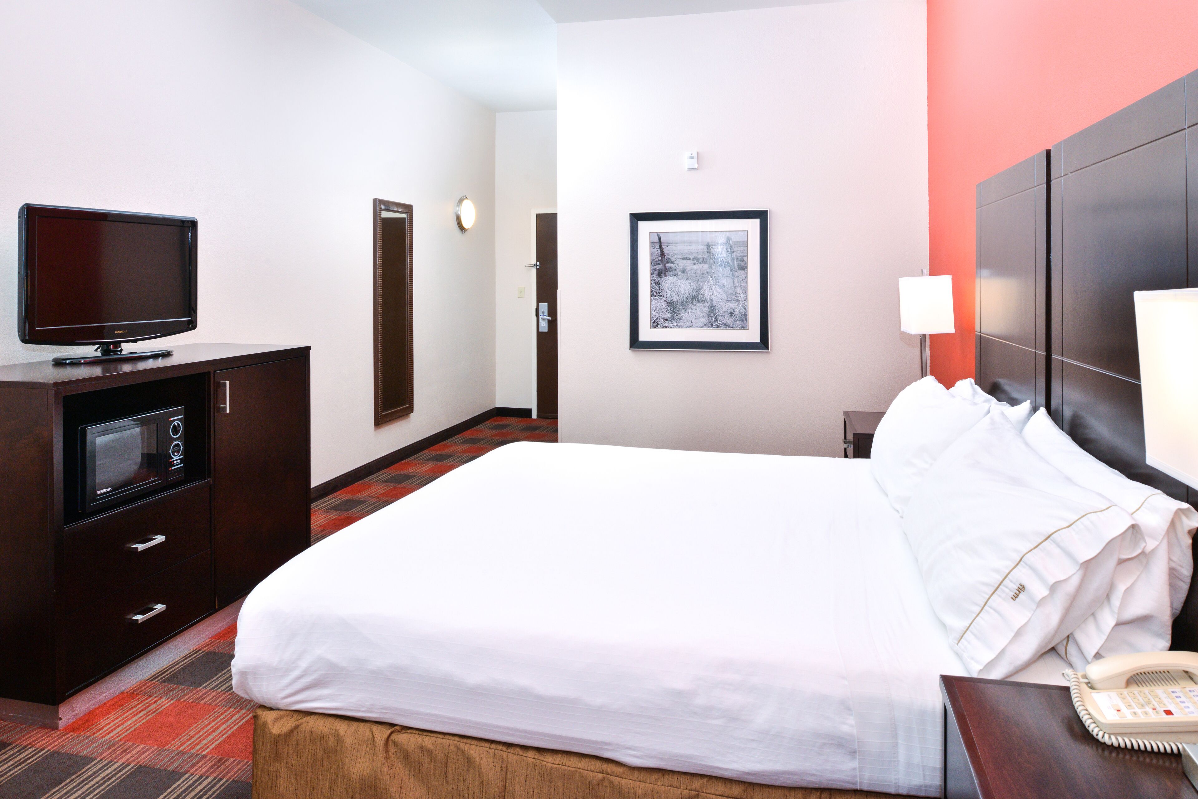 Une chambre d’hôtel avec un lit, un téléviseur posé sur une commode en bois, un tableau encadré au mur et une table de chevet avec une lampe et un téléphone.