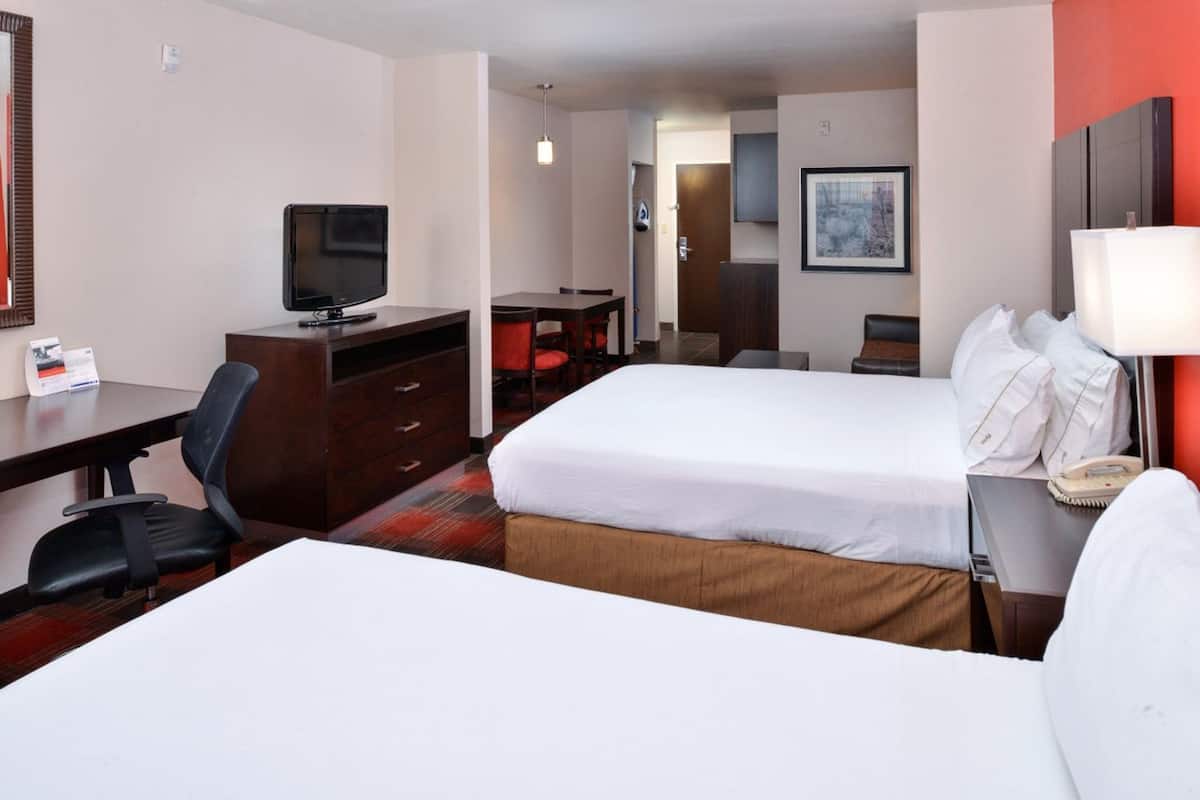 A hotel room with two beds, a desk with a television, a chair, and a bedside table with a lamp.
