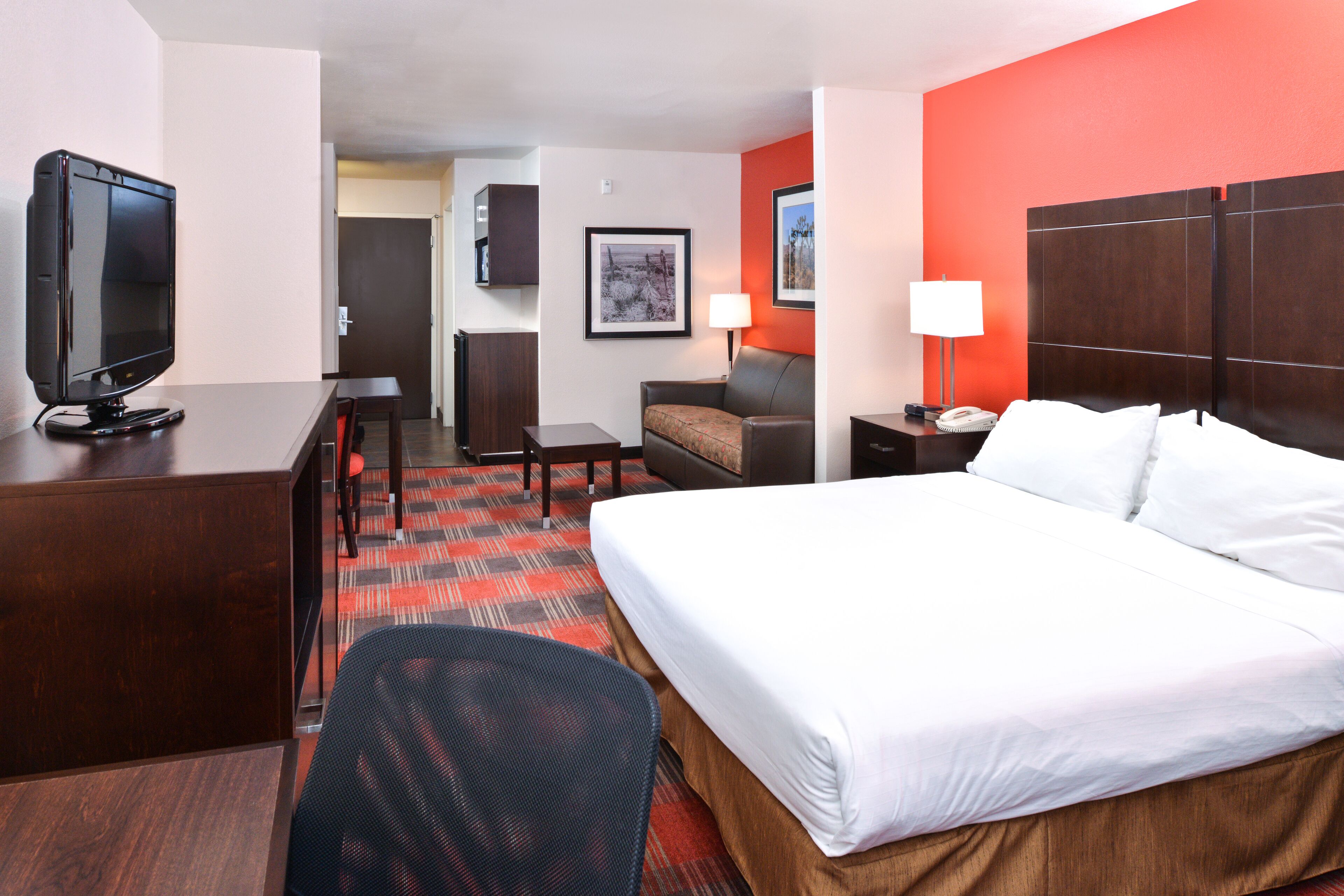 A hotel room with a bed, a desk with a television, a chair, a sofa, and a nightstand with a lamp.