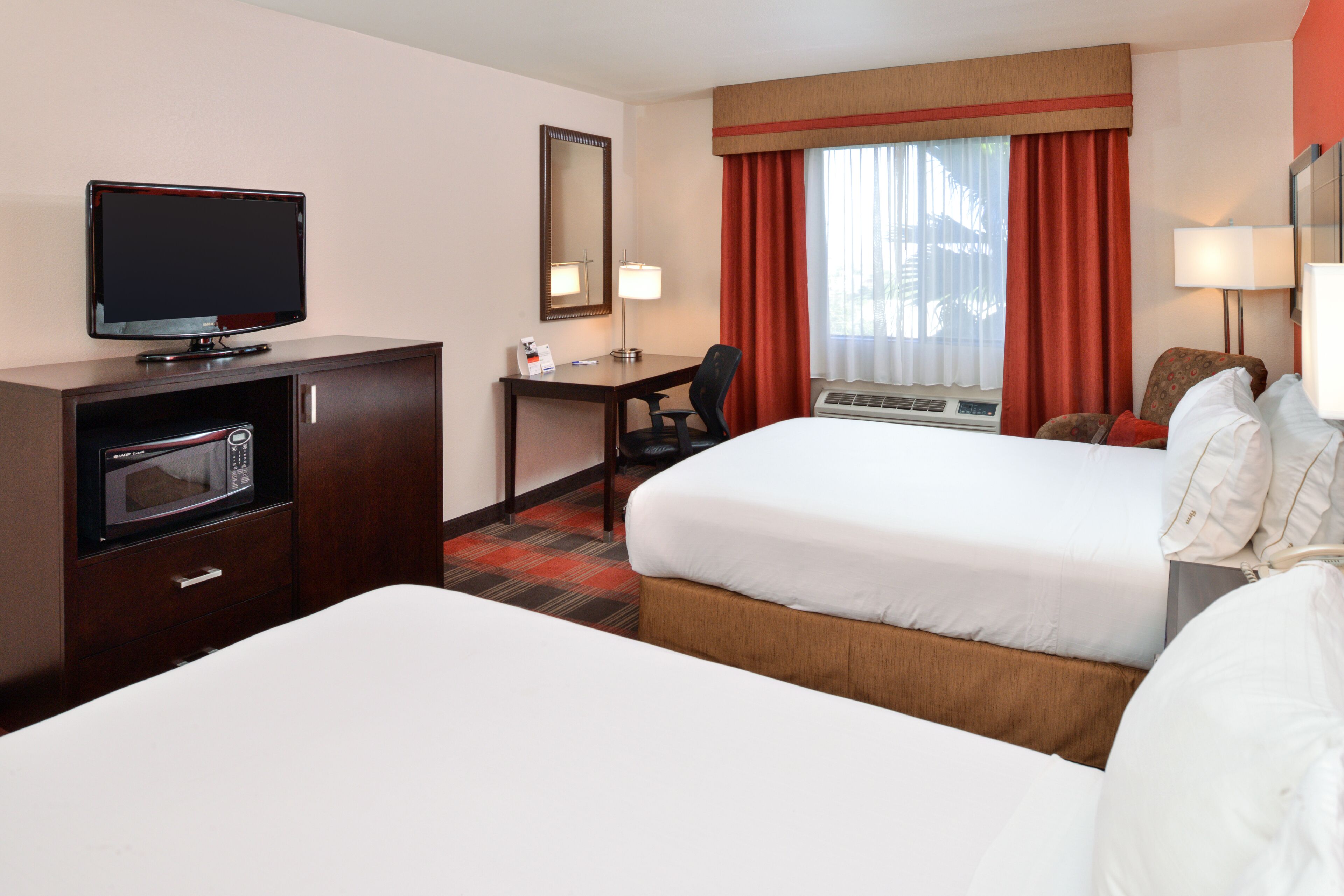 A hotel room with two beds, a desk with a chair, a television, a microwave, and a window with curtains.