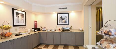 Una cocina de hotel con piso a cuadros, una cesta de frutas, una máquina de café y una zona de bufé con pastelería.