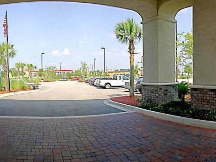 A paved entrance with a brick pathway, palm trees, and a white vehicle parked under a building with columns.