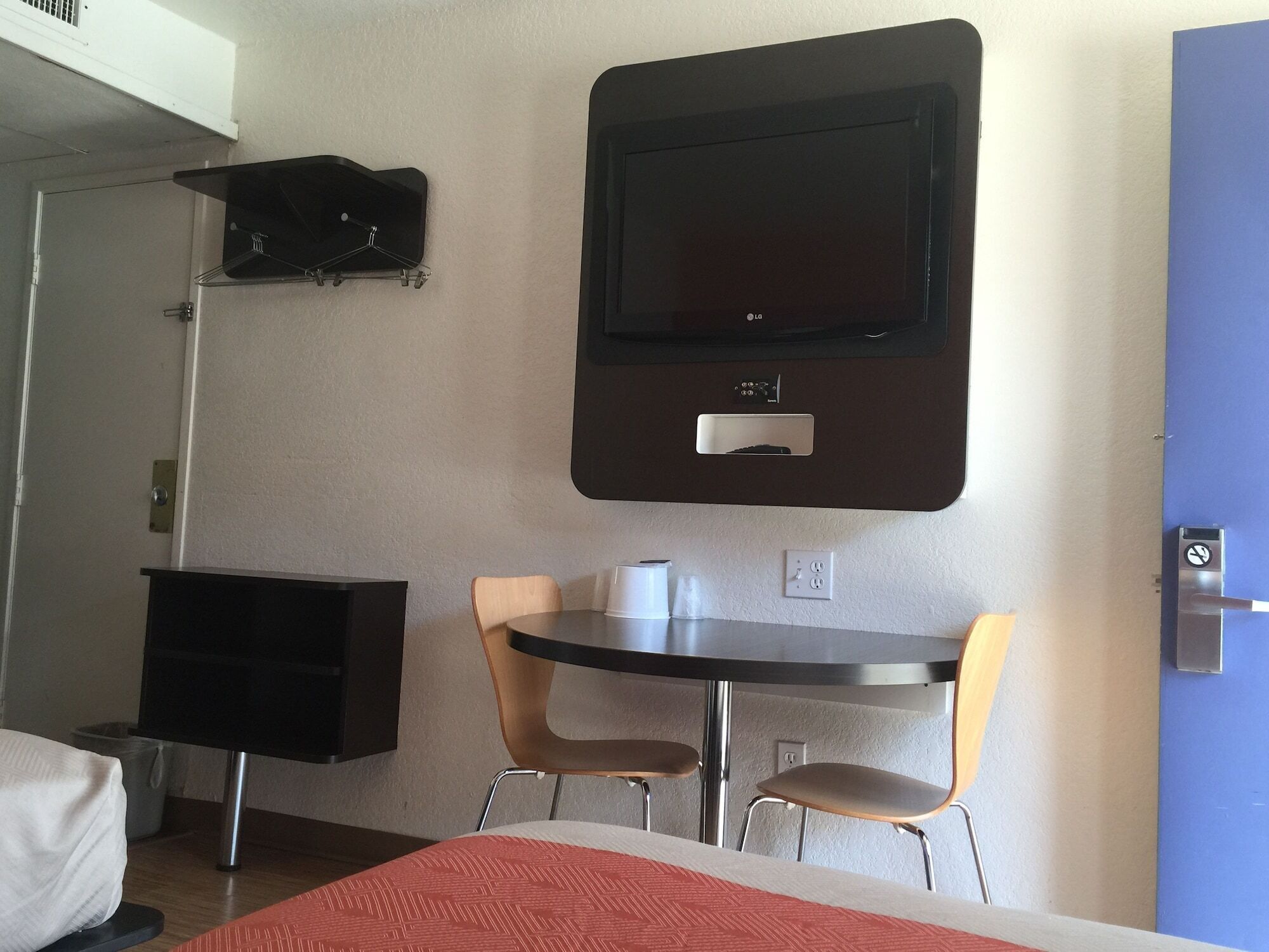 A hotel room with a bed, a desk with two chairs, a flat-screen TV mounted on the wall, and a coat rack.