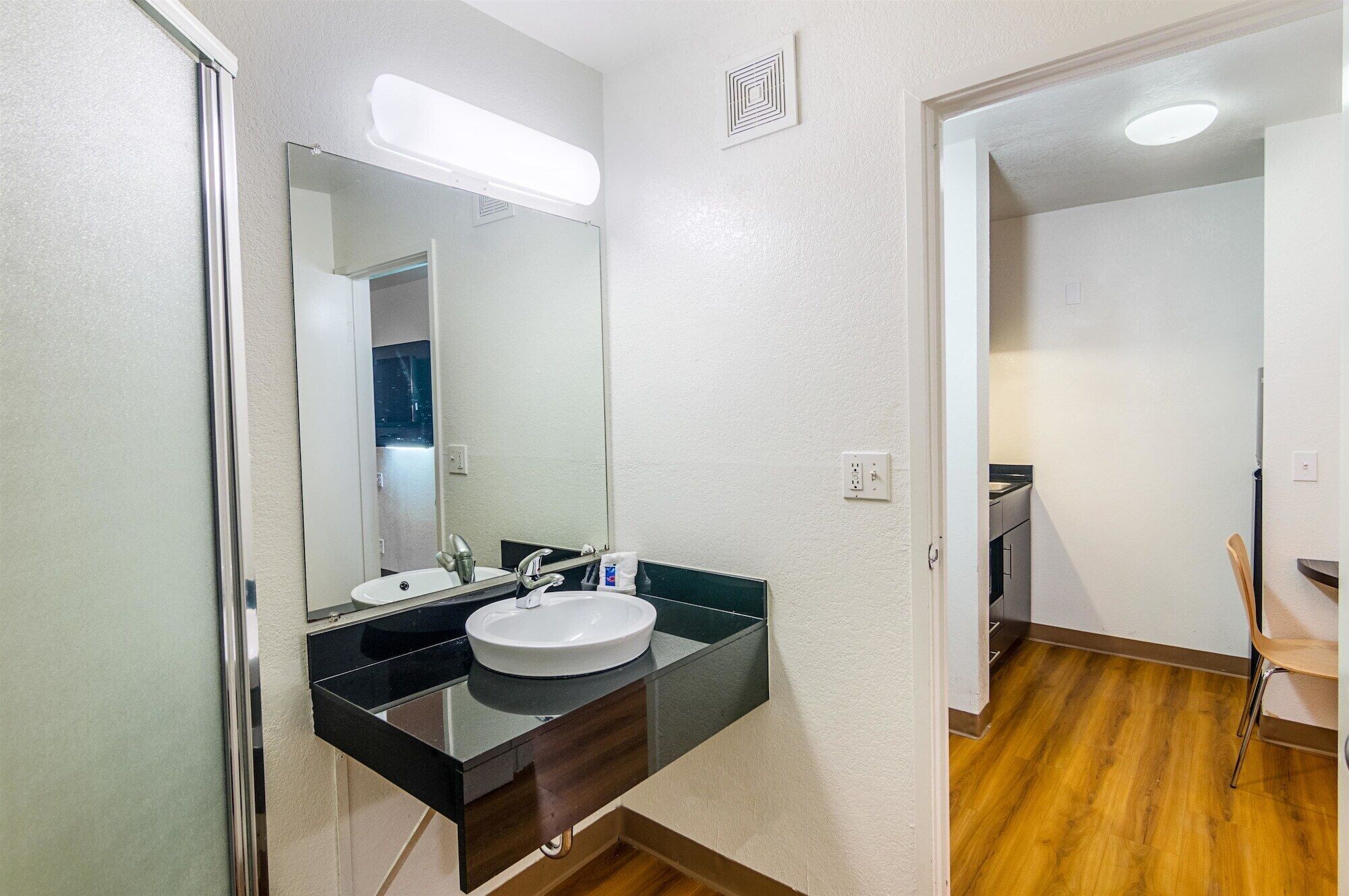 A bathroom with a white sink, black countertop, and a mirror. There is a wooden floor and a chair visible in the background.