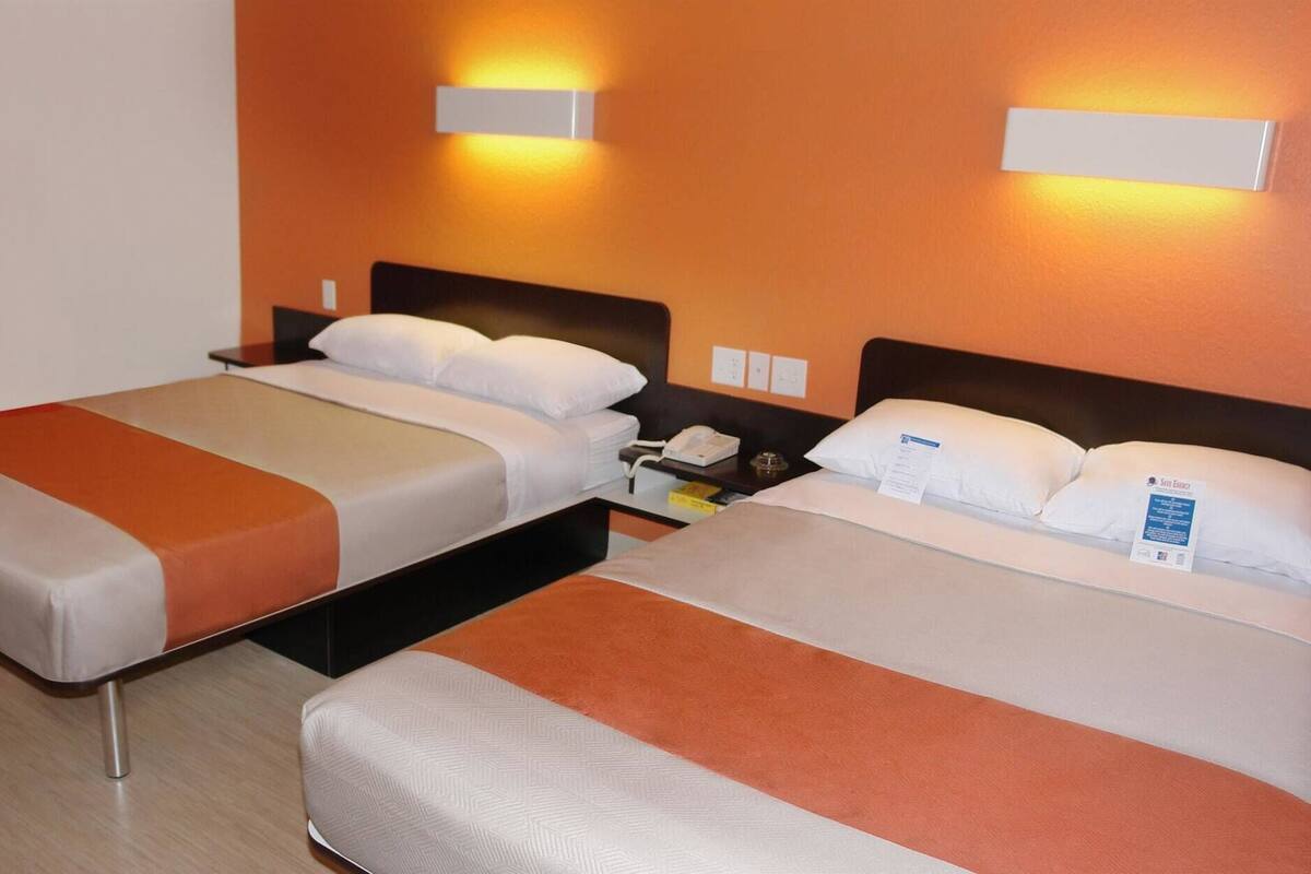A hotel room with two beds, each with a white and orange bedspread, a bedside table with a phone and a lamp, and wall-mounted light fixtures.