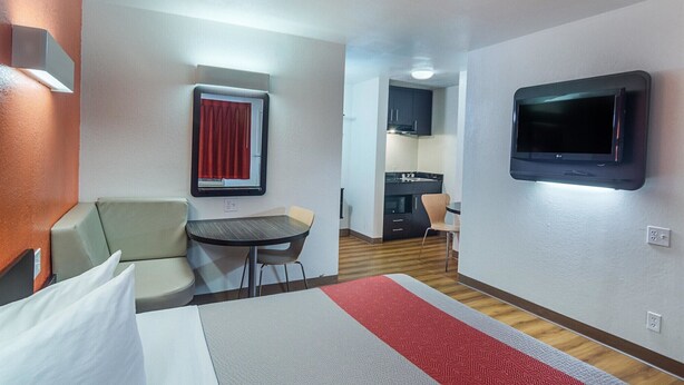 Une chambre d’hôtel compacte comprenant un lit, une petite table, un canapé, un téléviseur fixé au mur, une kitchenette et une fenêtre avec un rideau rouge.