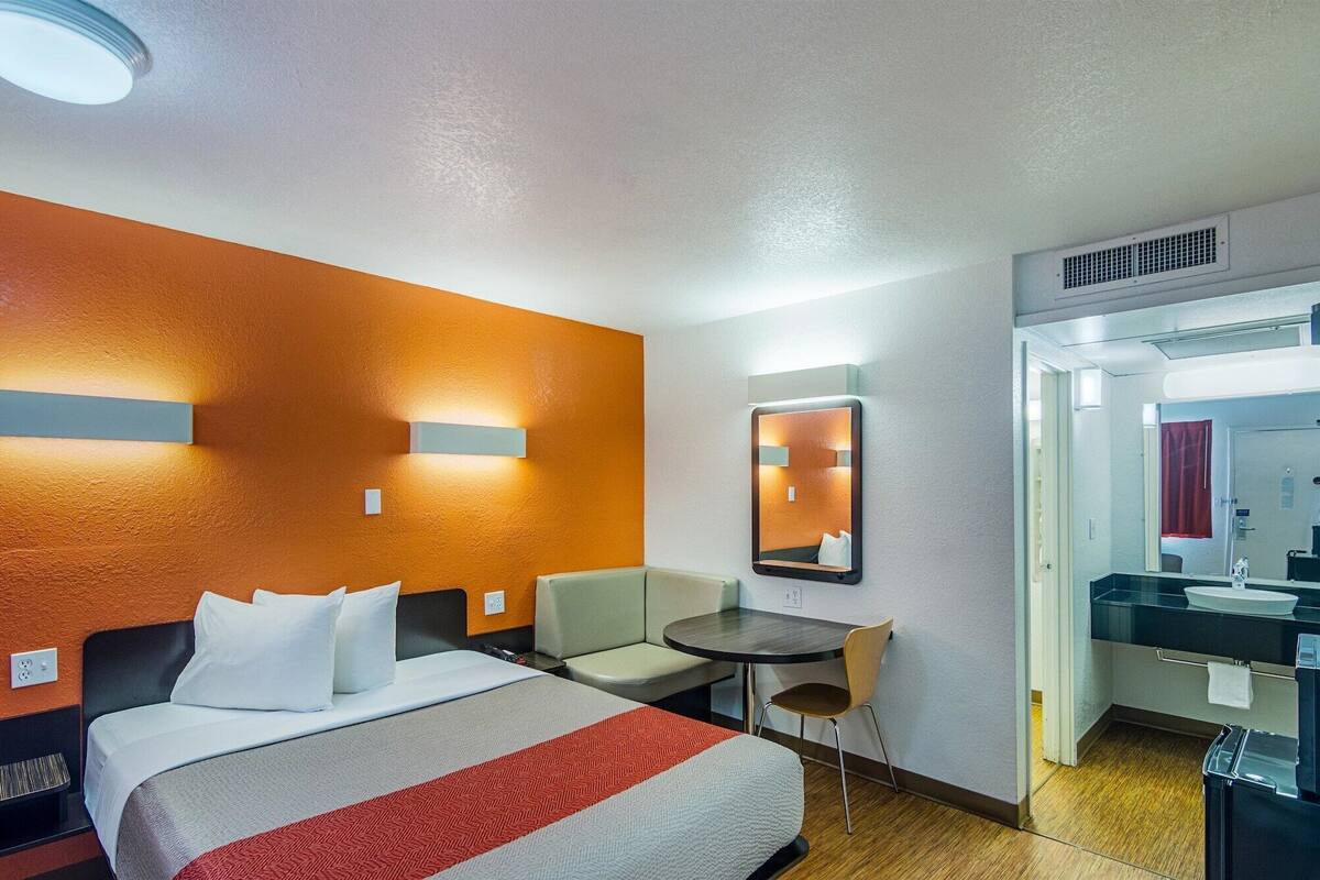 A hotel room with a bed, a small table, a chair, a mirror, and a bathroom visible through an open door.