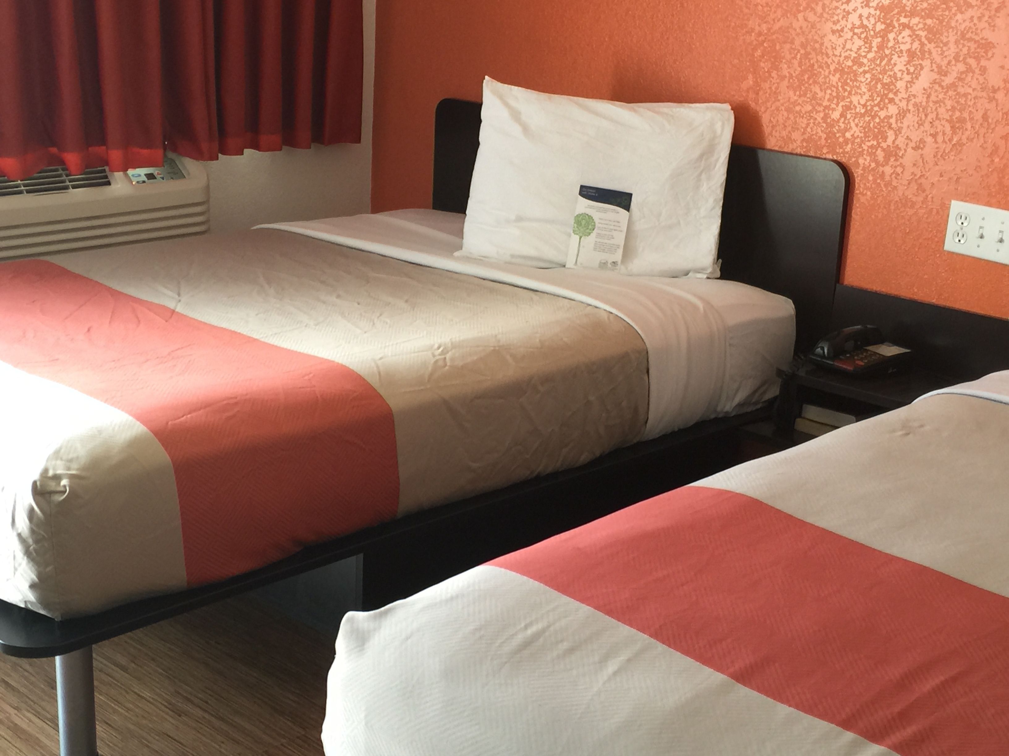 Habitación de hotel con dos camas, cada una con una colcha blanca y naranja, un cojín blanco y una mesita de noche con un teléfono y un libro.