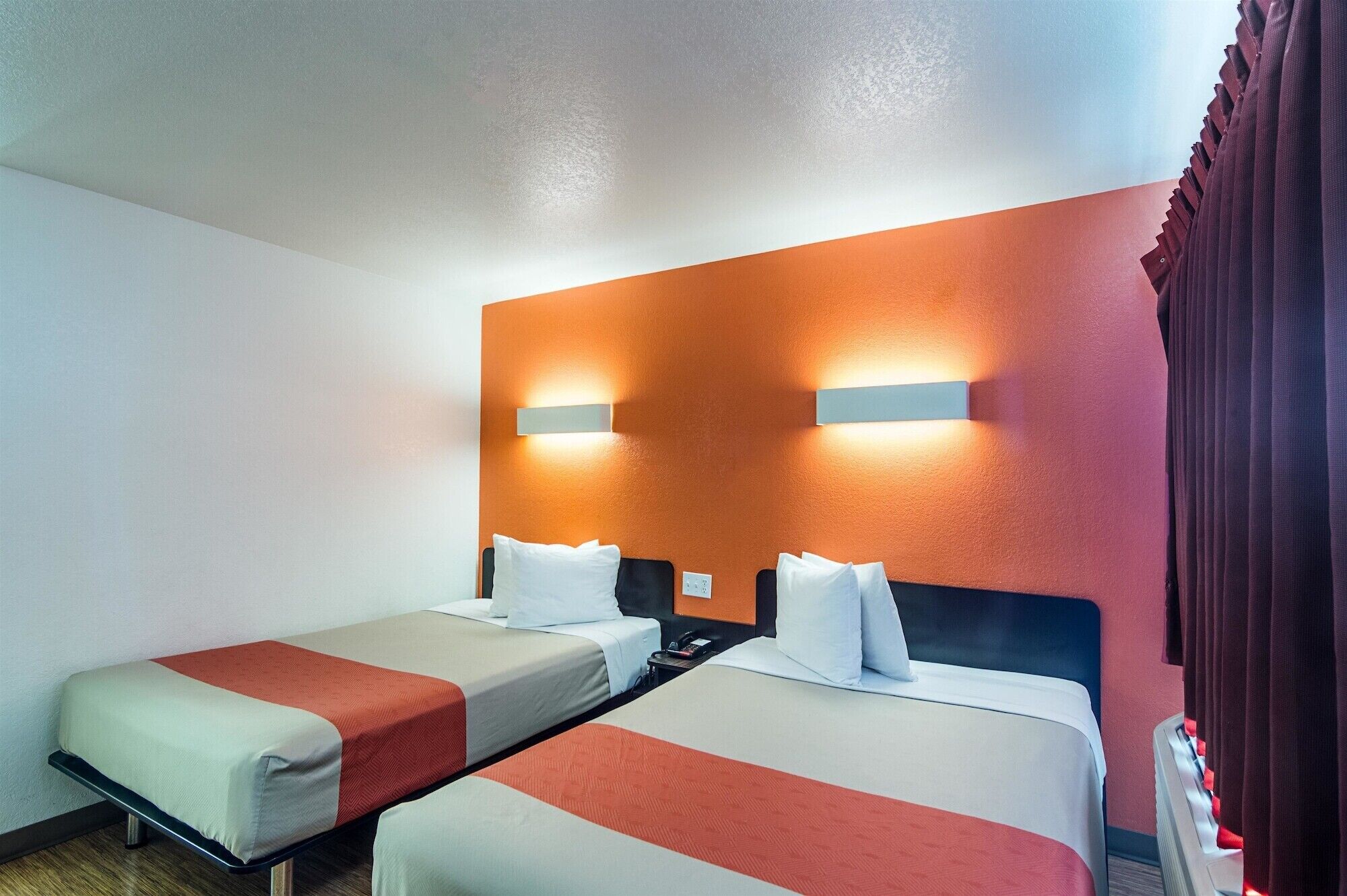 A hotel room with two single beds, a red-orange accent wall, and two wall-mounted light fixtures.