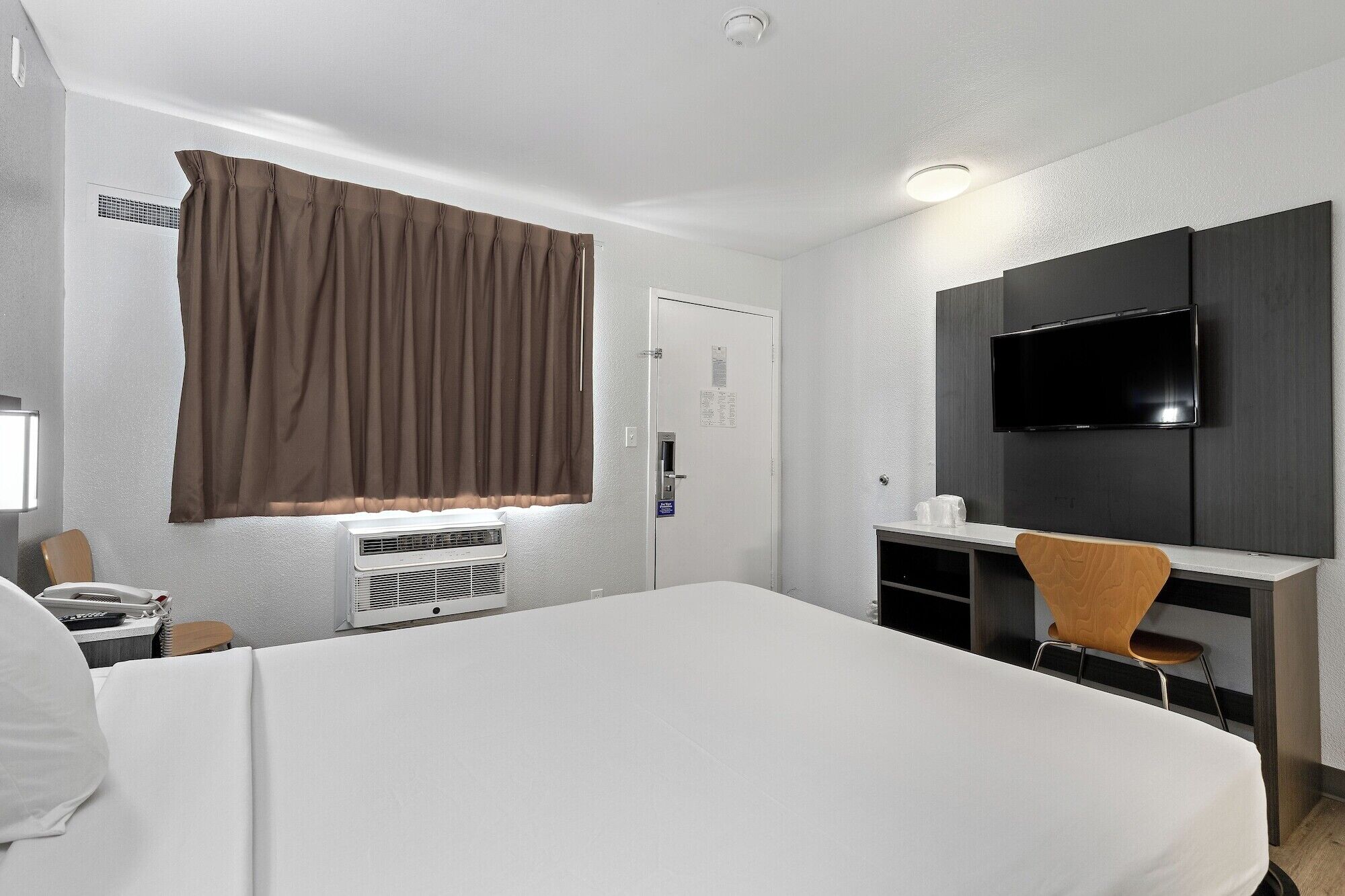 A hotel room with a large bed, a desk with a chair, a television, and an air conditioning unit.