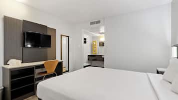 A modern hotel room with a large bed, a desk with a flat-screen TV, a chair, and a small kitchenette.