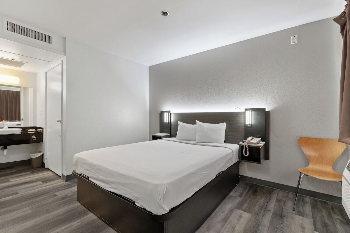 Habitación de hotel con una cama grande, una mesita de noche con un teléfono, una silla y un baño visible a través de una puerta abierta.