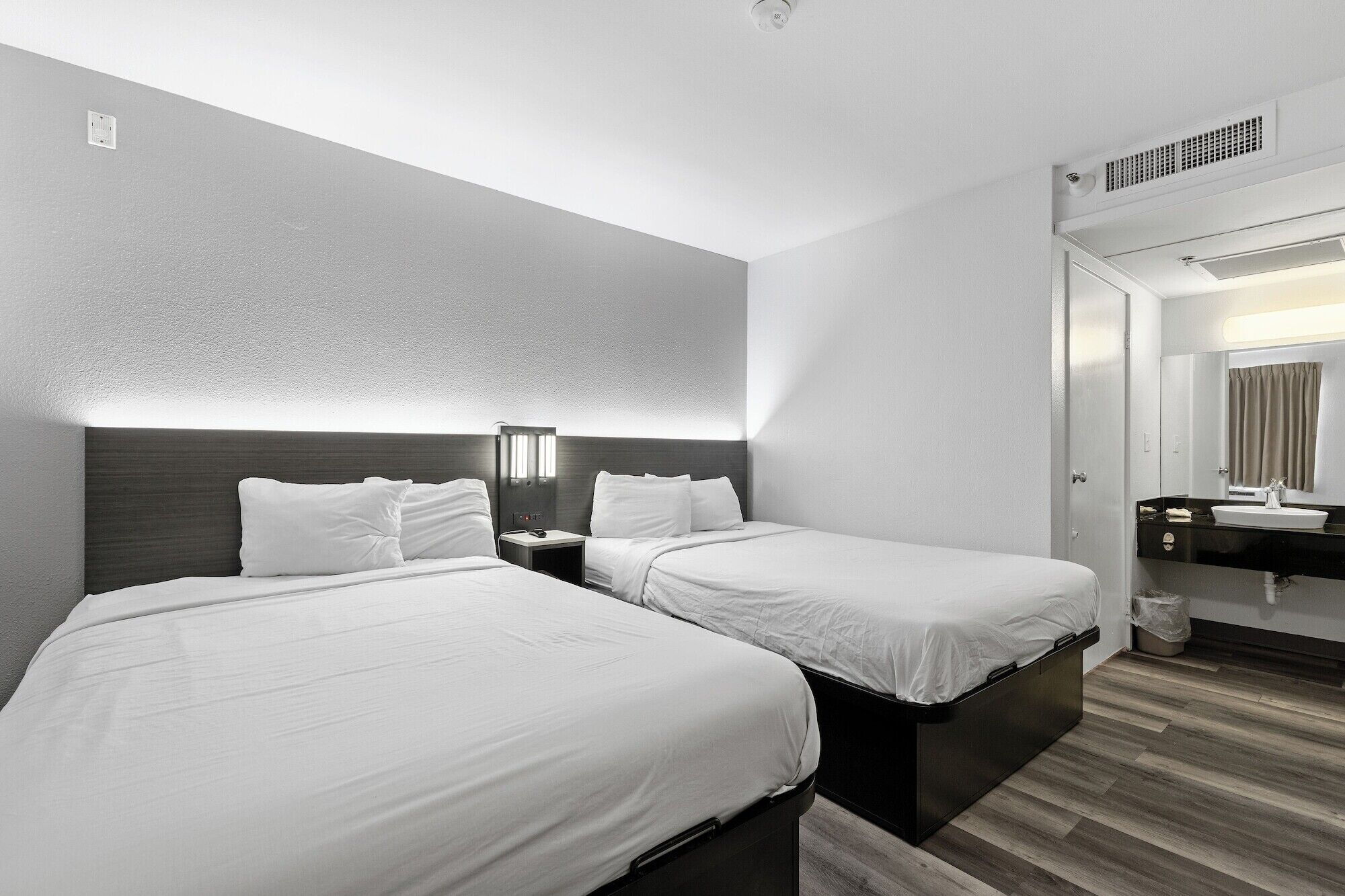 A hotel room with two beds, a nightstand, a bathroom with a sink and mirror, and a ceiling vent.
