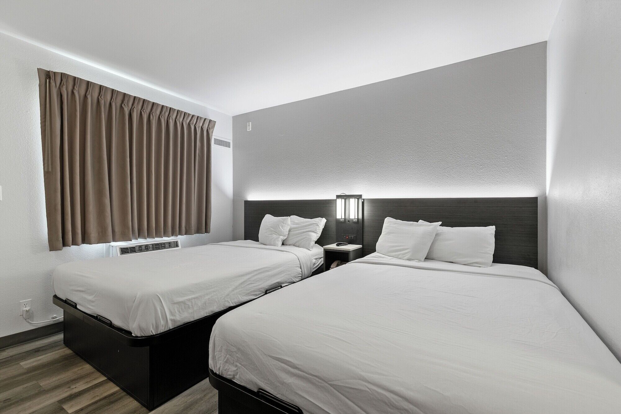 A hotel room with two single beds, a nightstand with a lamp, a window with curtains, and a wall-mounted air conditioning unit.