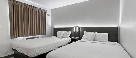 A hotel room with two single beds, a nightstand with a lamp, a window with curtains, and a wall-mounted air conditioning unit.