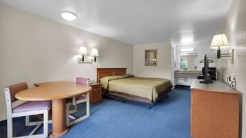 A hotel room with a bed, a desk, a chair, a small table, and a lamp.