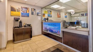 A reception area with a digital screen displaying a message, a coffee machine, and a desk with a computer. - Motel 6 Villa Park, IL - Chicago West (Villa Park)