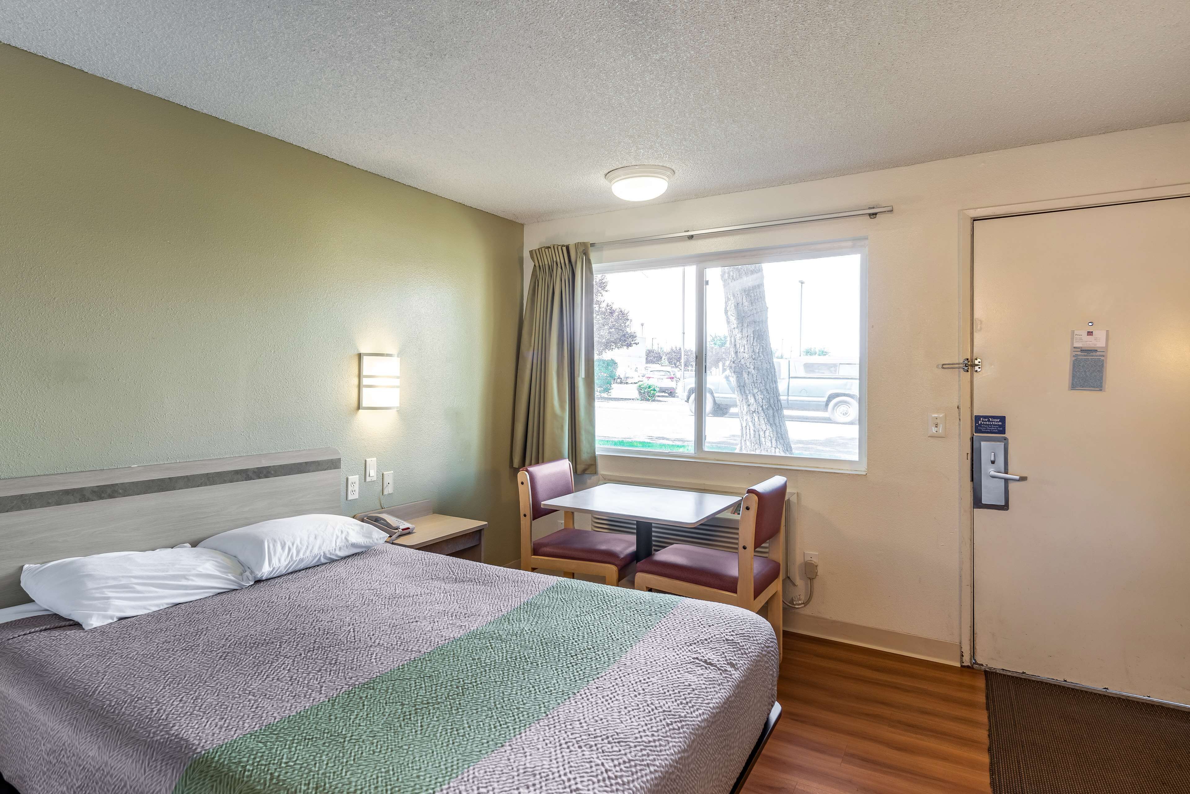 Quarto de hotel com uma cama, uma pequena mesa com cadeiras, uma janela com cortinas e uma porta com fechadura.