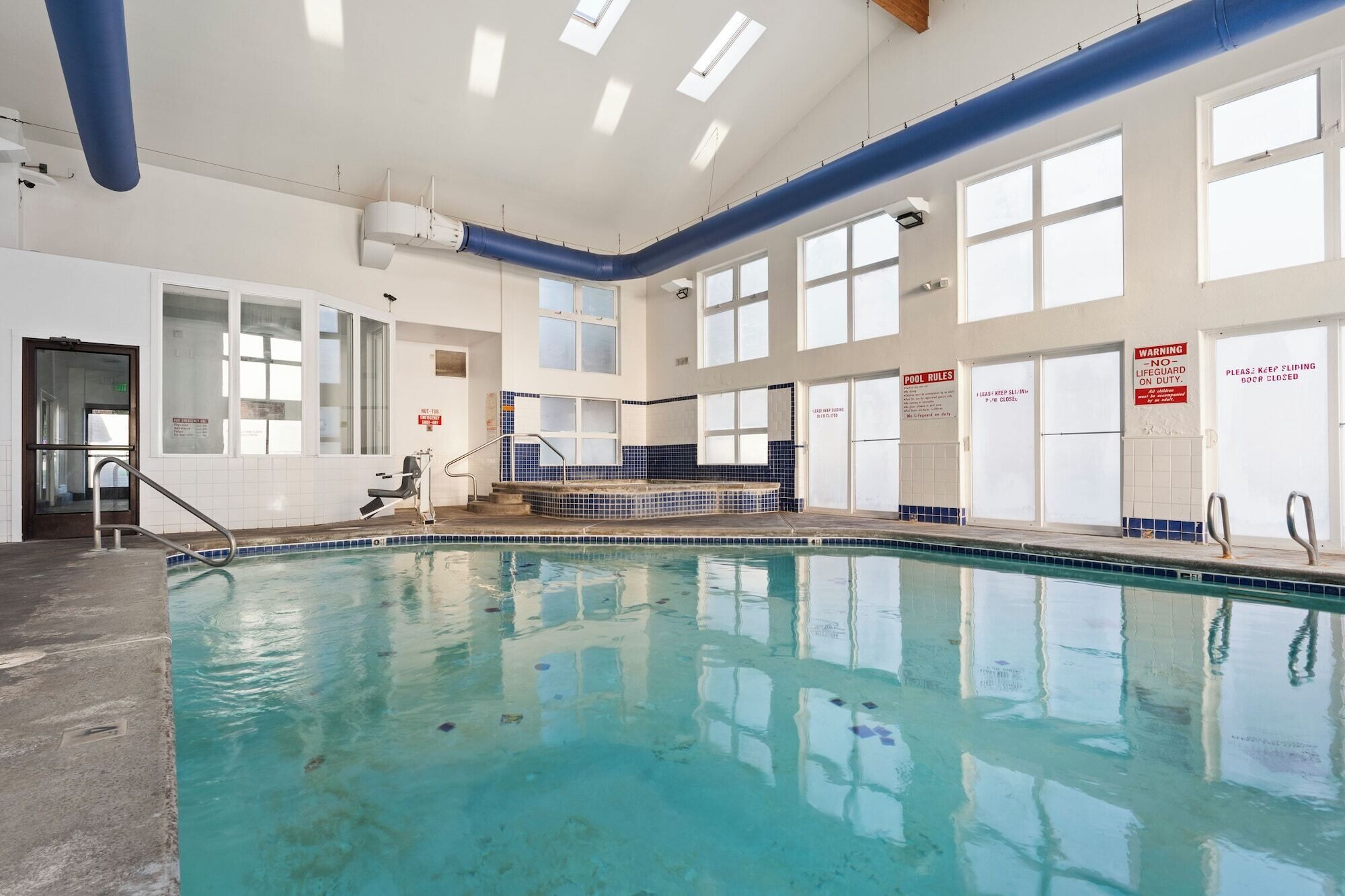 An indoor swimming pool with clear blue water, large windows, and safety signs.